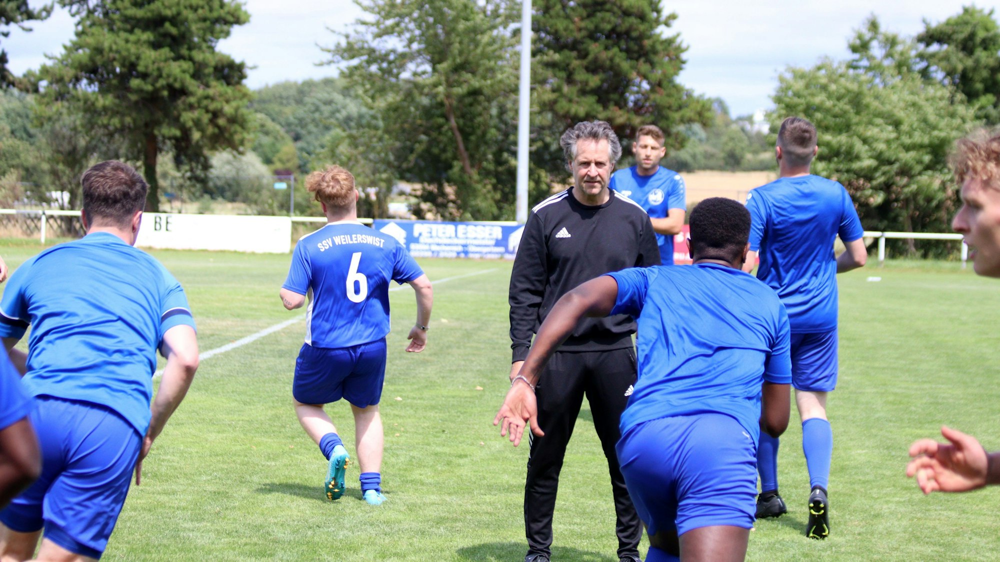 Trainer Horst Bartz steht auf dem Fußballplatz und beobachtet seine Spieler, die Laufübungen absolvieren.
