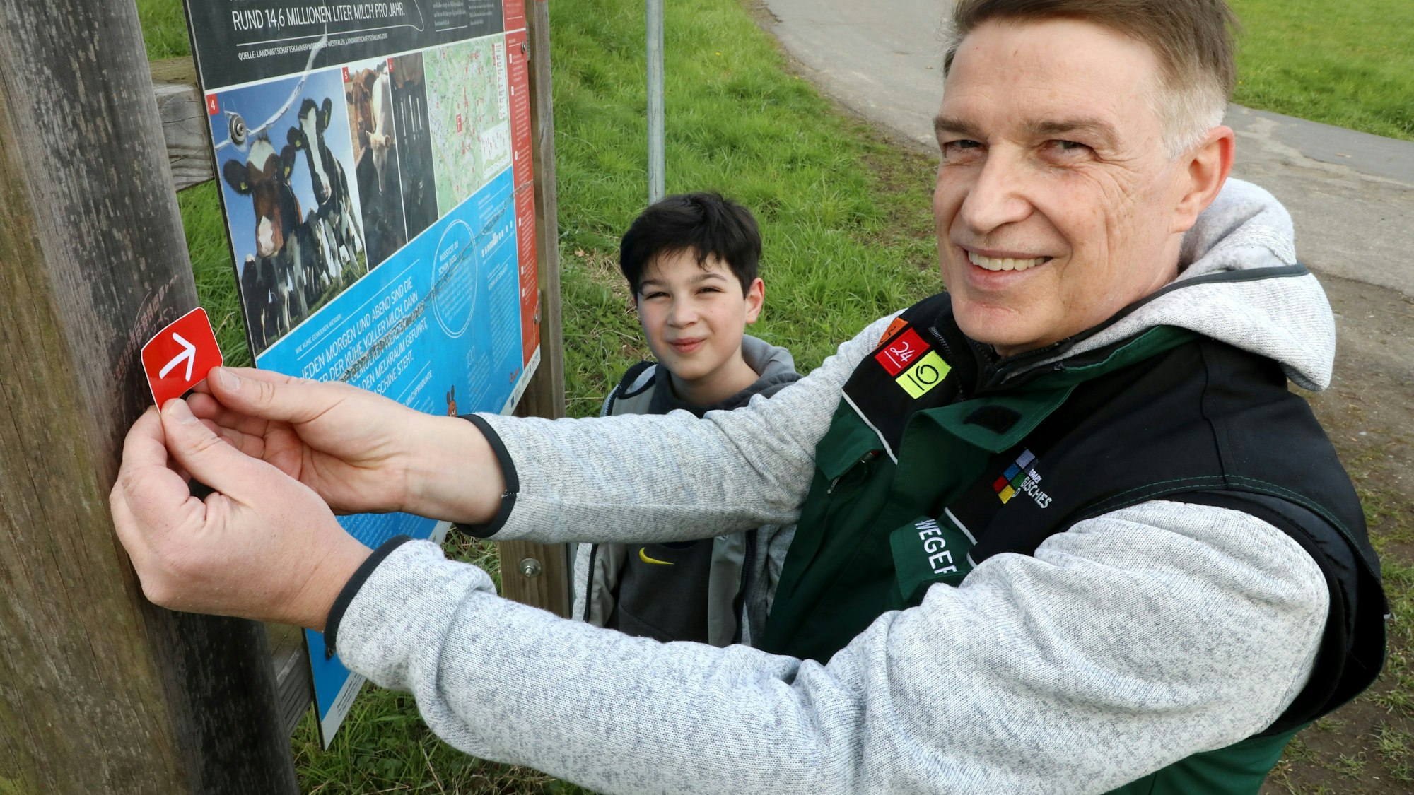Dr. Erik Werdel klebt als Wegepate des Bauernhofwegs in Lohmar eine Markierung an einen Pfosten. Im Hintergrund ist sein Sohn Maximilian zu sehen.