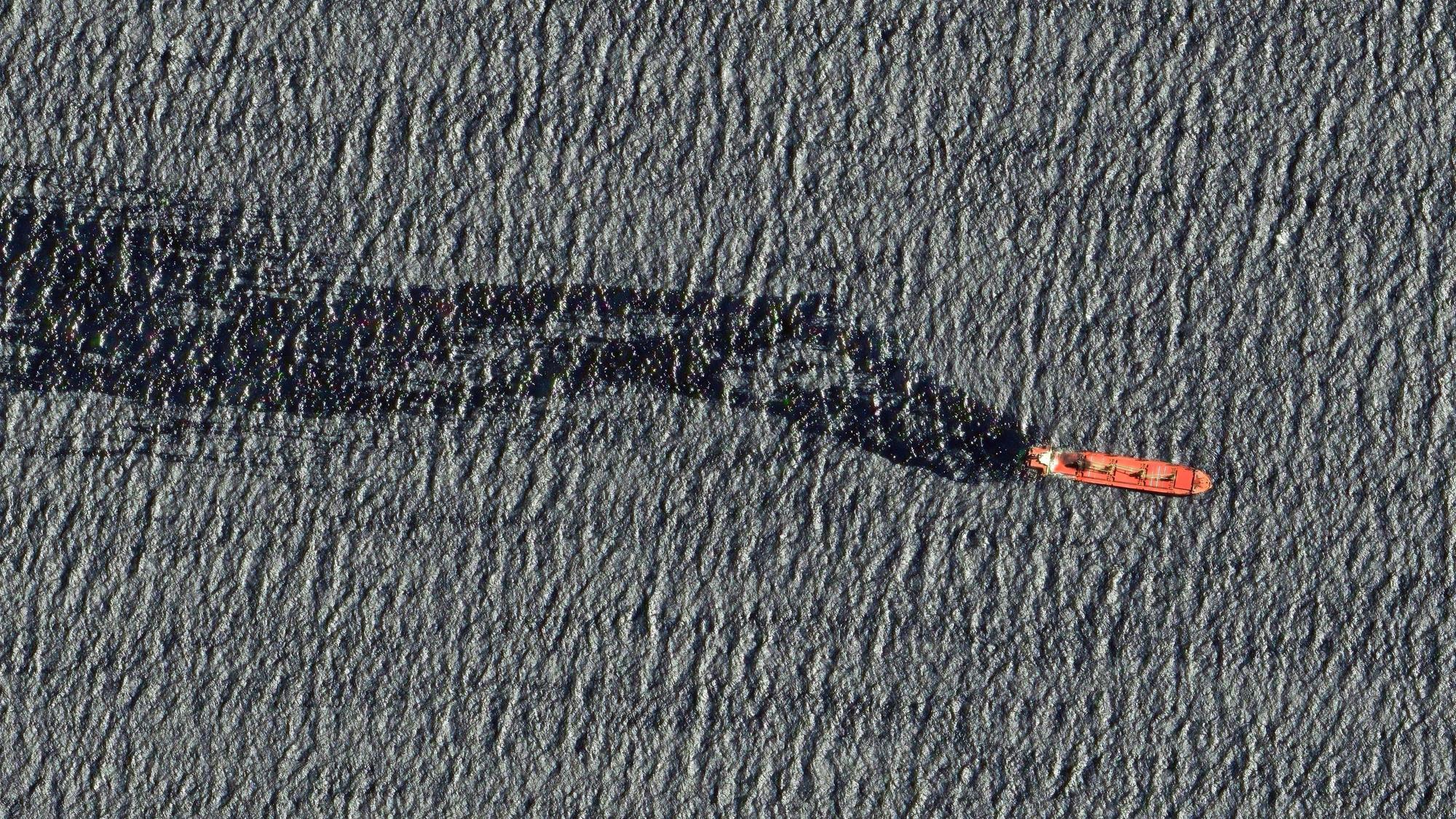 Bild von oben: Ein rotes Schiff fährt auf dem Meer. Der Frachter hinterlässt einen großen schwarzen Ölteppich hinter sich.