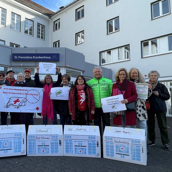 Menschen stehen vor dem Eingang des St. Franziskus-Krankenhaus Eitorf, vor ihnen Häuschen mit den Ortsnamen der bereits geschlossenen Geburtsstationen in der Region.