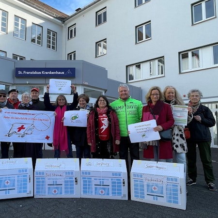 Menschen stehen vor dem Eingang des St. Franziskus-Krankenhaus Eitorf, vor ihnen Häuschen mit den Ortsnamen der bereits geschlossenen Geburtsstationen in der Region.