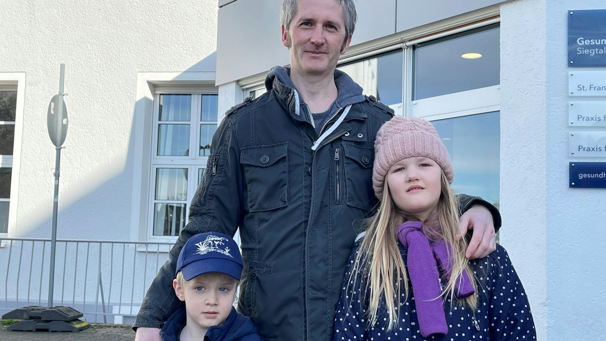 Ein Mann steht mit einem blonden Jungen und einem langhaarigen Mädchen vor dem Eingang des St. Franziskus-Krankenhauses in Eitorf.