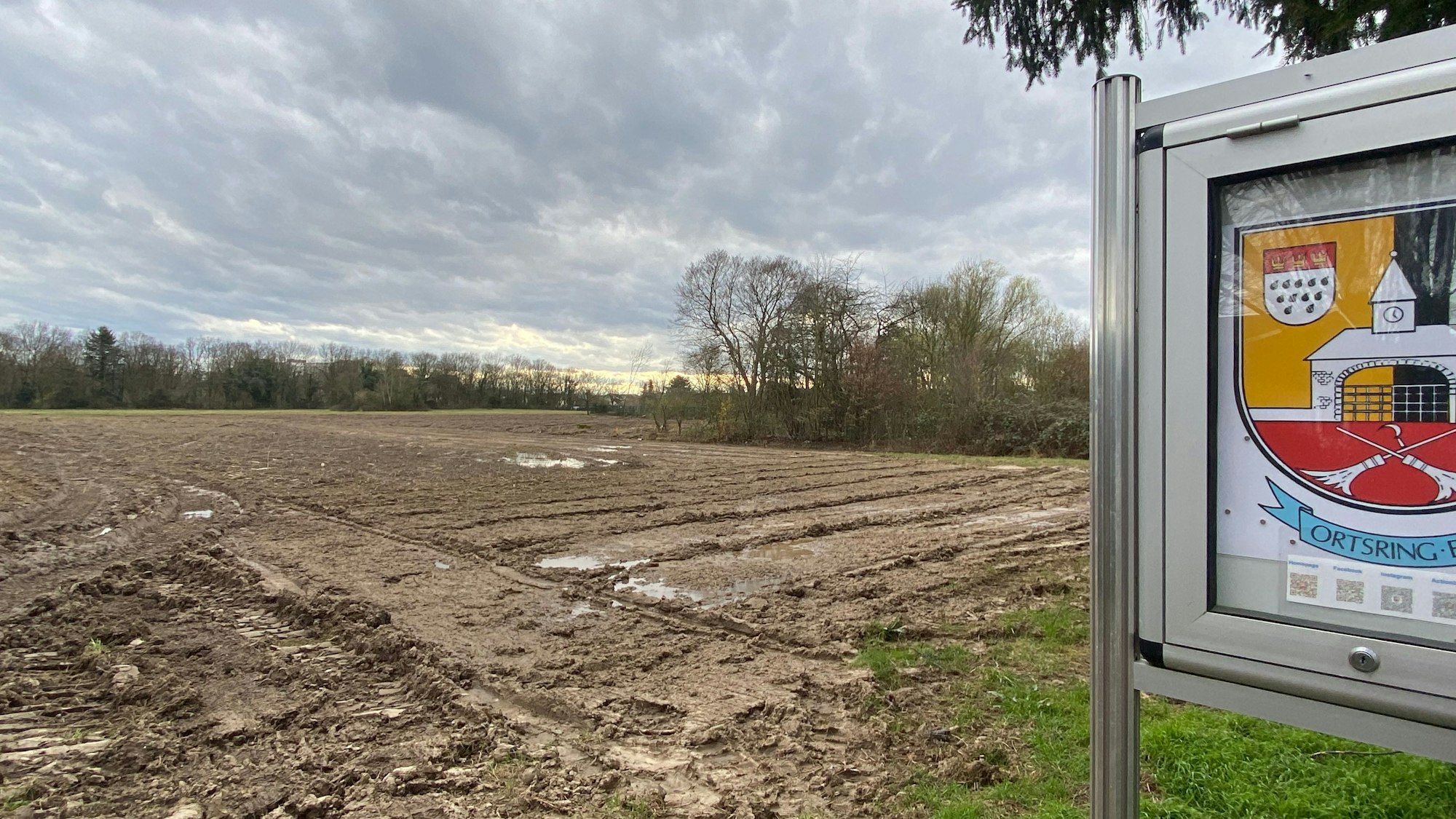 Blick auf einen Acker, in der Ferne sind Bäume und am rechten Bildrand ein Schaukasten des Ortsrings Eil zu sehen.