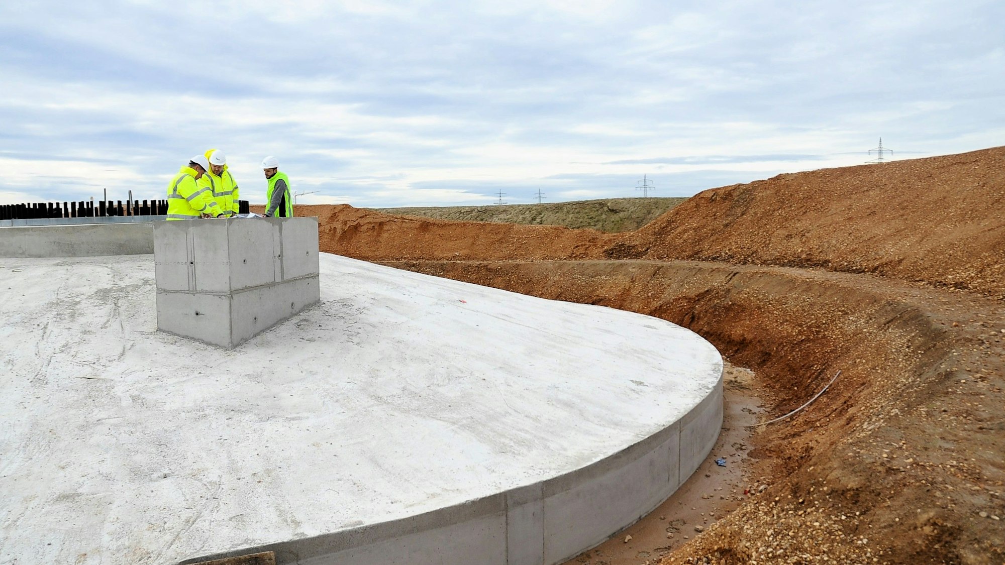 Zu sehen sind drei Männer in Warnwesten, die auf einem Fundament für ein Windrad stehen.