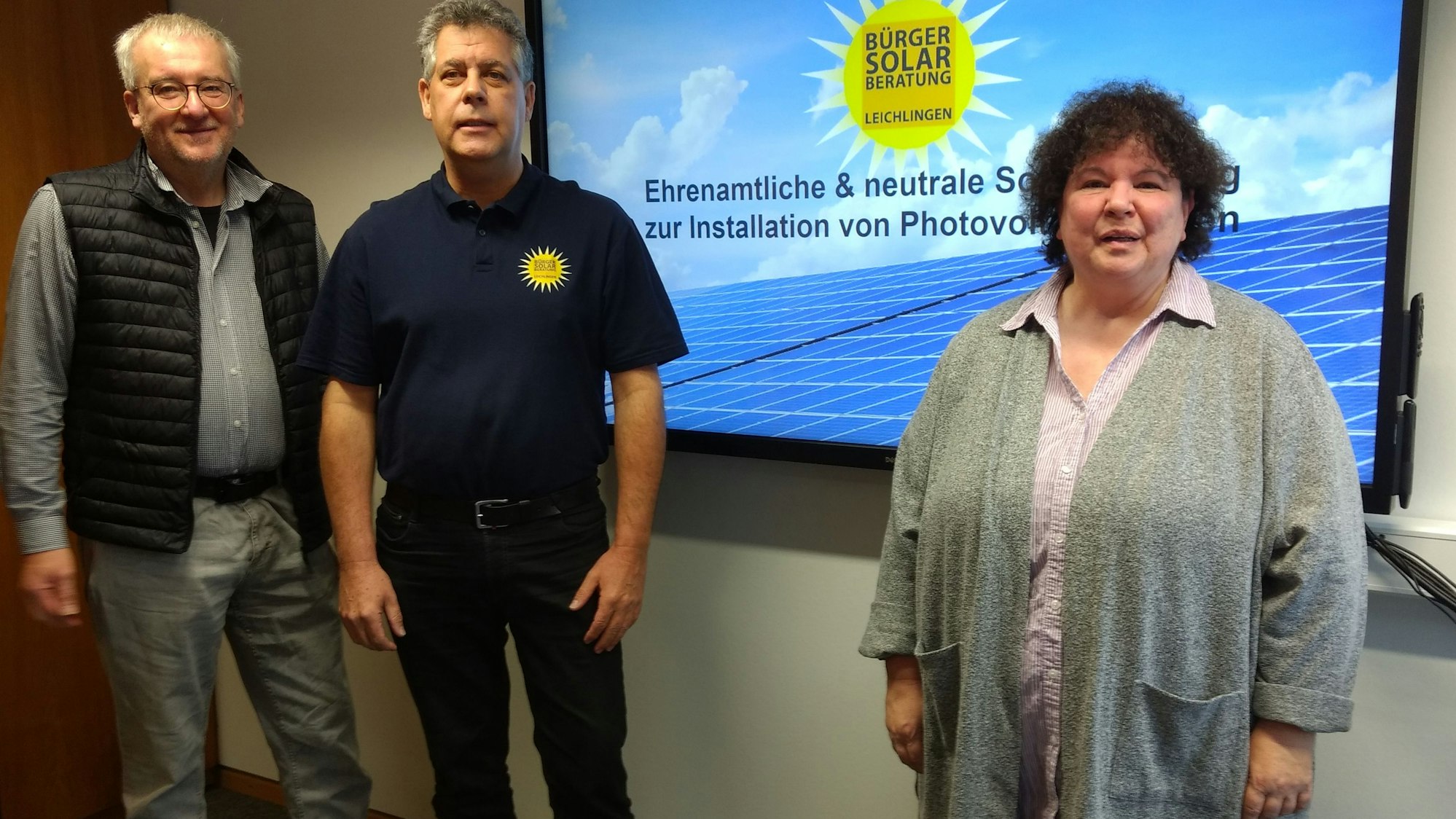 Zwei Männer und eine Frau stehen vor einem Bildschirm.