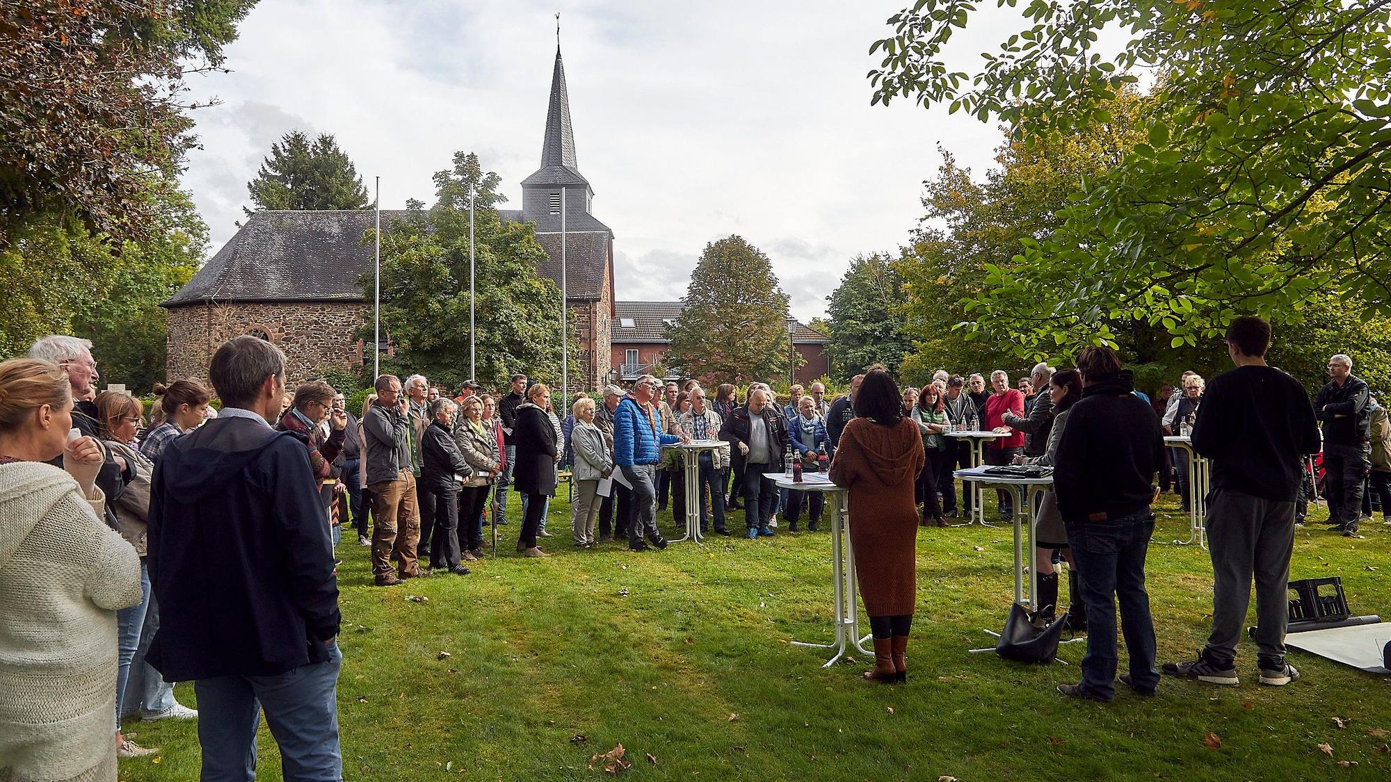 Zahlreiche Menschen stehen bei einer Bürgerversammlung in Heimbach-Blens auf einer Wiese, im Hintergrund ist die Kirche zu sehen.
