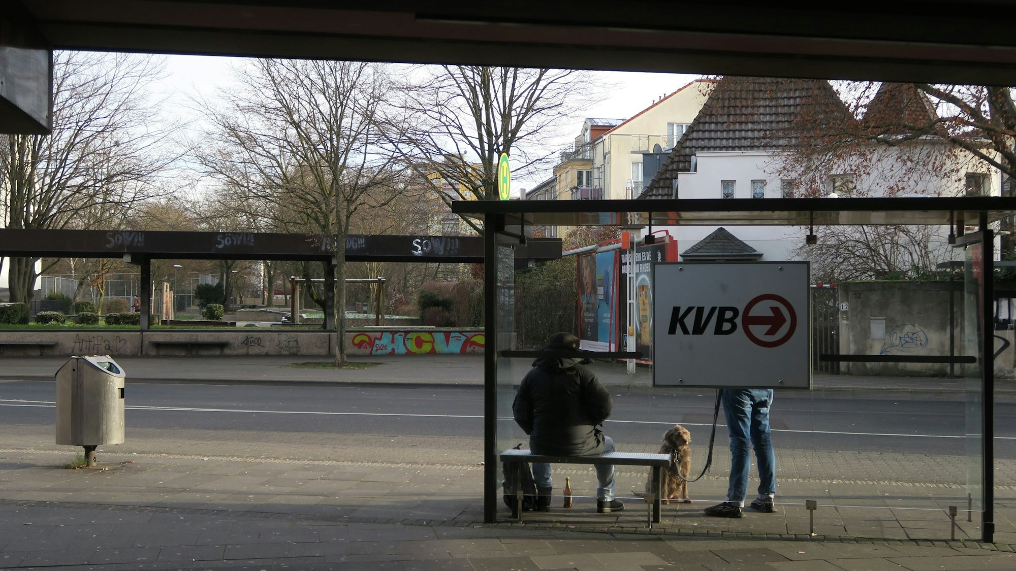 An einer Bushaltestelle warten zwei Fahrgäste, einer hat einen Hund dabei.