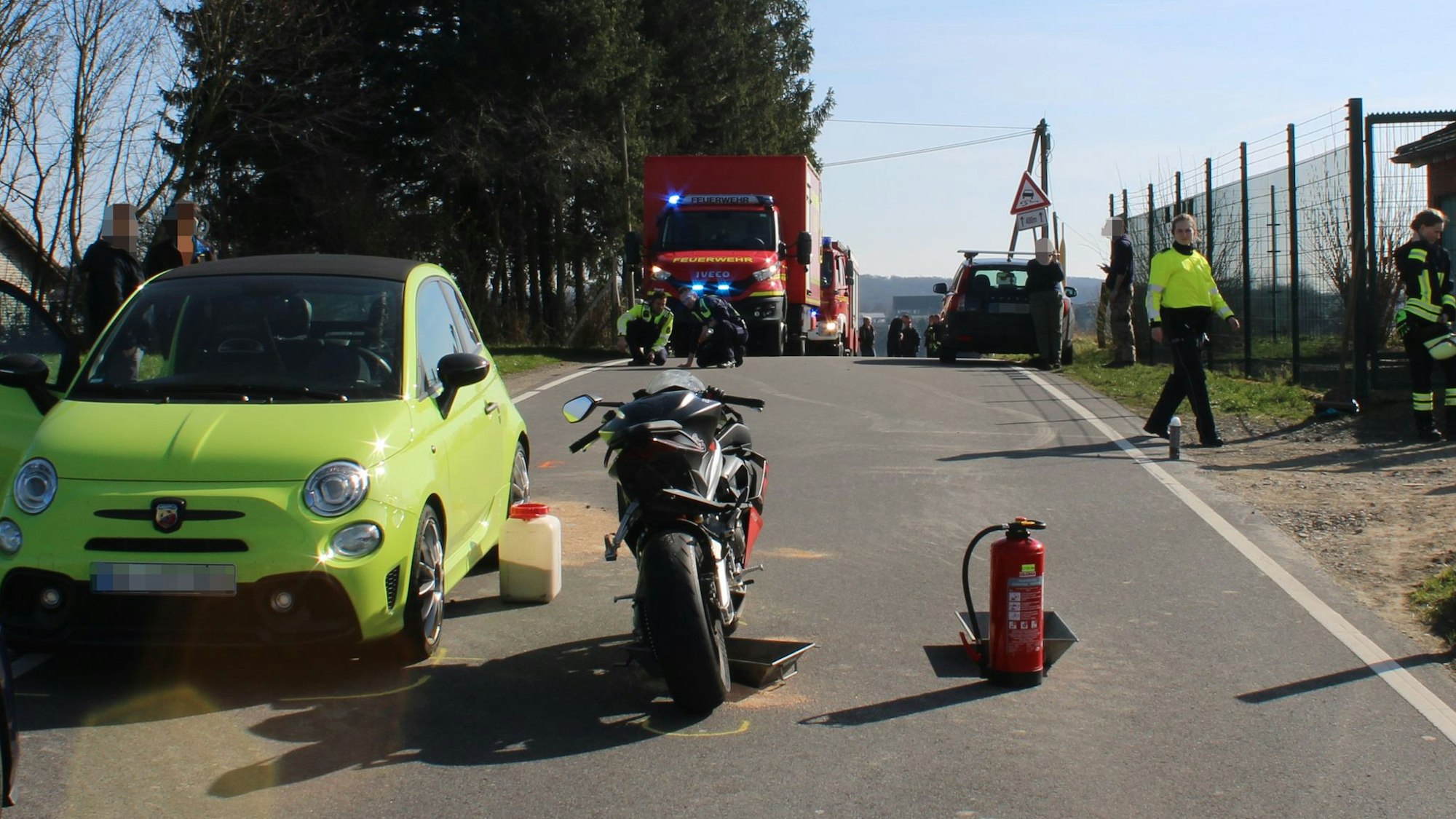 Beschädigte Zweiräder und ein Auto stehen auf einer Fahrbahn, auf dem Boden ist Ölbindemittel zu sehen. Im Hintergrund stehen Einsatzkräfte der Feuerwehr.
