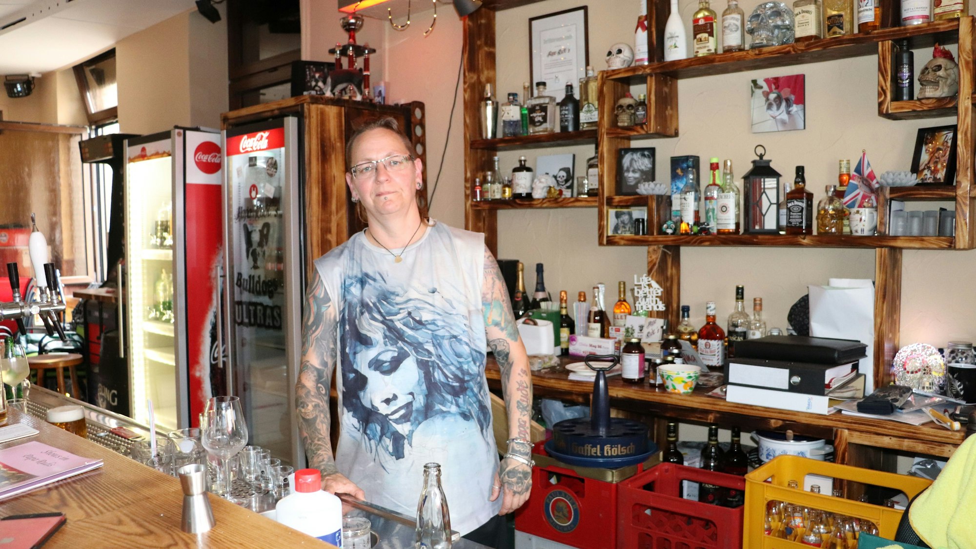 Eine Frau mit Tina-Turner-Shirt und tätowierten Armen steht hinter der Theke einer Kneipe.