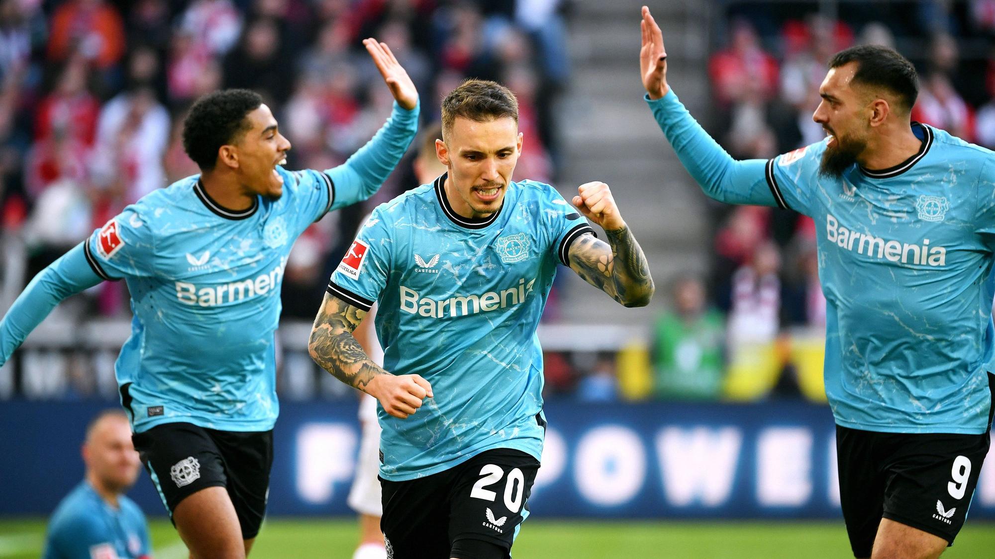 Alejandro Grimaldo (m.) jubelt nach seinem Tor zum 0:2 gegen den 1. FC Köln. Hinter ihm sind Amine Adli (l.) und Borja Iglesias (r.) im Begriff, mit Händen einzuschlagen.