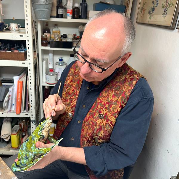 Ein Mann mit Glatze und Brille sitzt mit Schürze an einem Arbeitstisch und hantiert mit einer Porzellanfigur.
