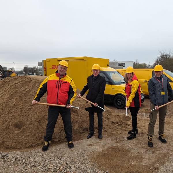 Vier Männer und eine Frau mit gelben Helmen und Spaten auf der Baustelle. Im Hintergrund gelbe Zustellfahrzeuge der Post.