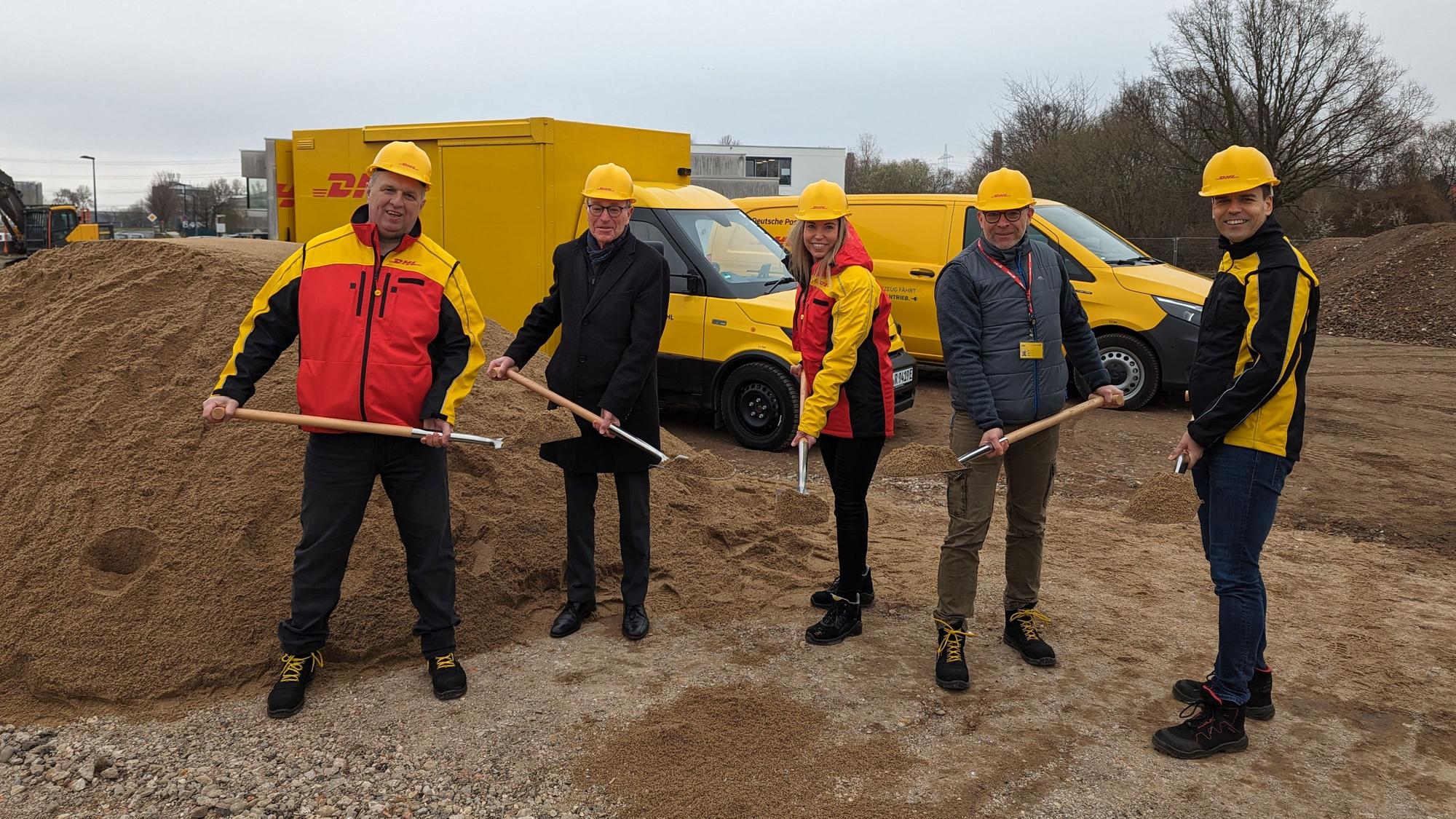 Vier Männer und eine Frau mit gelben Helmen und Spaten auf der Baustelle. Im Hintergrund gelbe Zustellfahrzeuge der Post.