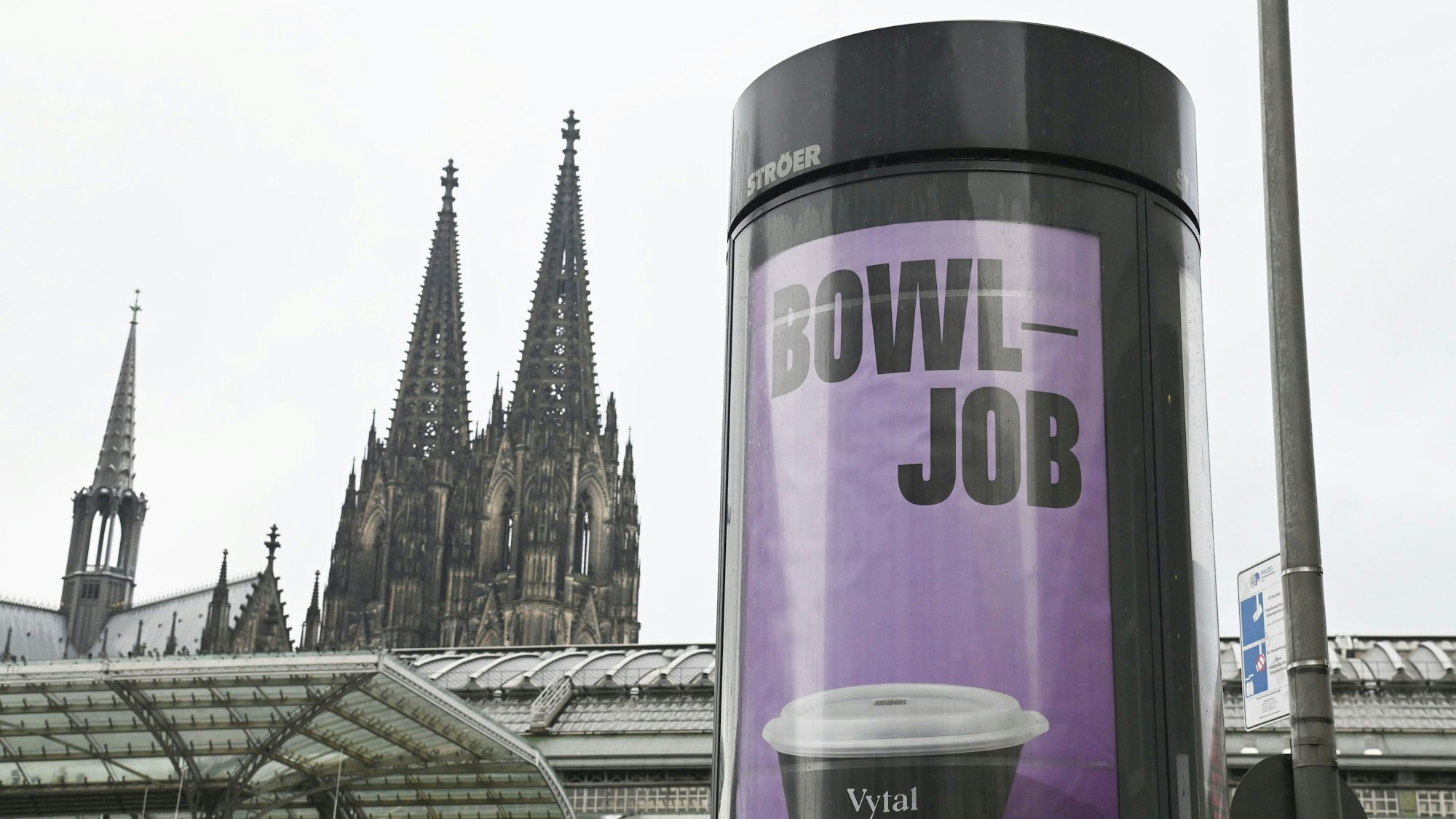 Plakate auf einer Litfaßsäule mit dem Kölner Dom im Hintergrund: Das Kölner Unternehmen Vytal warb mit dem Kunstwort „Bowljob“ für seine Mehrwegbehälter, auf die nicht direkt Pfand gezahlt werden muss.