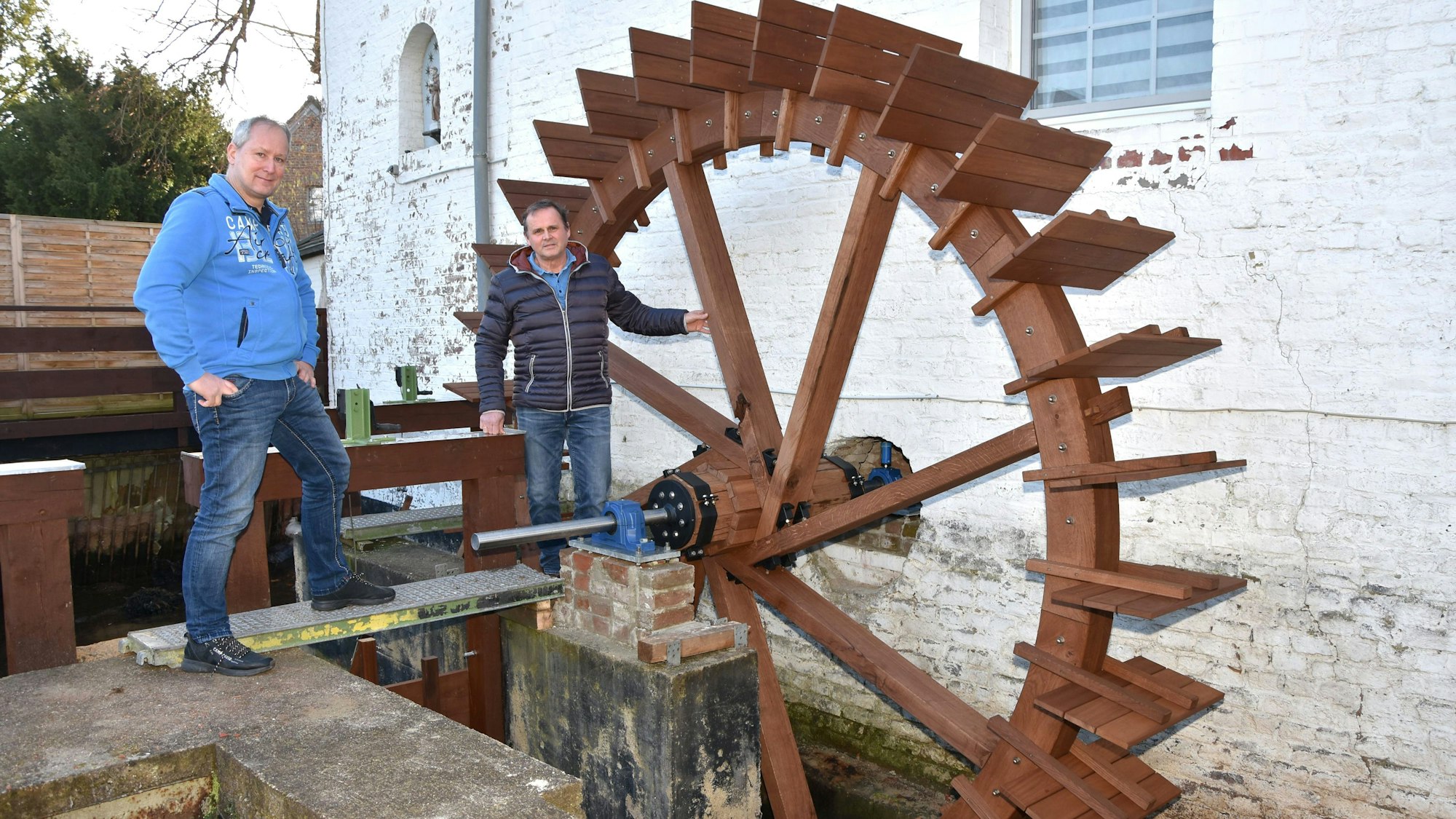 Zwei Männer stehen an einem hölzernen Mühlrad einer Wassermühle.