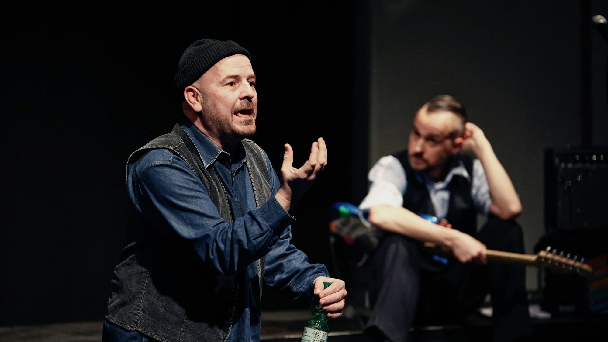 Manuel Moser steht im Vordergrund und gestikuliert stark beim Sprechen. Er trägt eine schwarze Weste und eine schwarze Mütze. Im Hintergrund sitzt Tim Mrosek mit einer E-Gitarre auf dem Schoß und stützt sich den Kopf mit der Hand.