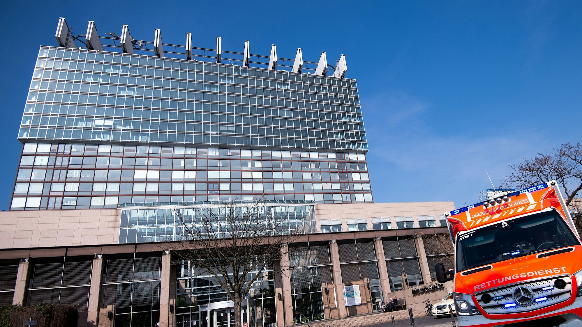 Zu sehen sind das Bettenhaus der Kölner Uniklinik und ein Rettungswagen in der Vorderansicht.