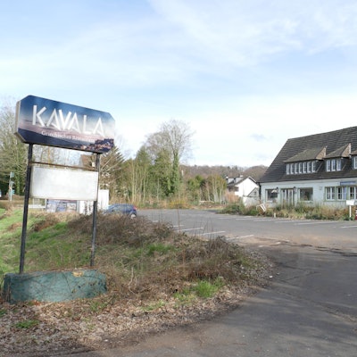 Das geschlossene Restaurant Kavala in Lohmar-Donrath: Ein weißes Haus mit Spitzdach und gauben, davor ein leerer Parkplatz. Unkraut wächst und Müll liegt an der Treppe.