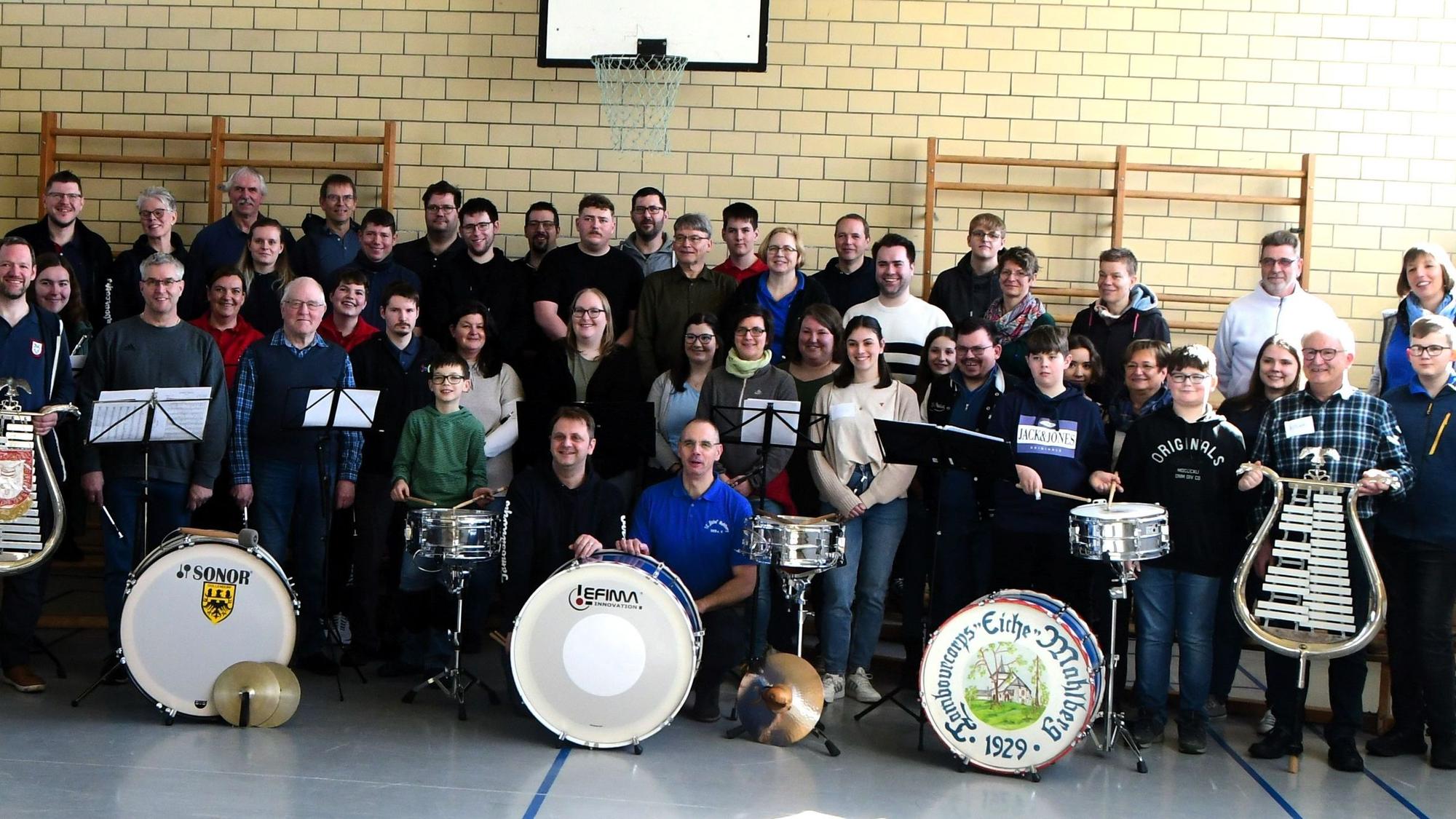 Zahlreiche Musiker stehen in einer Turnhalle. Einige halten Lyras, vor anderen stehen Pauken.