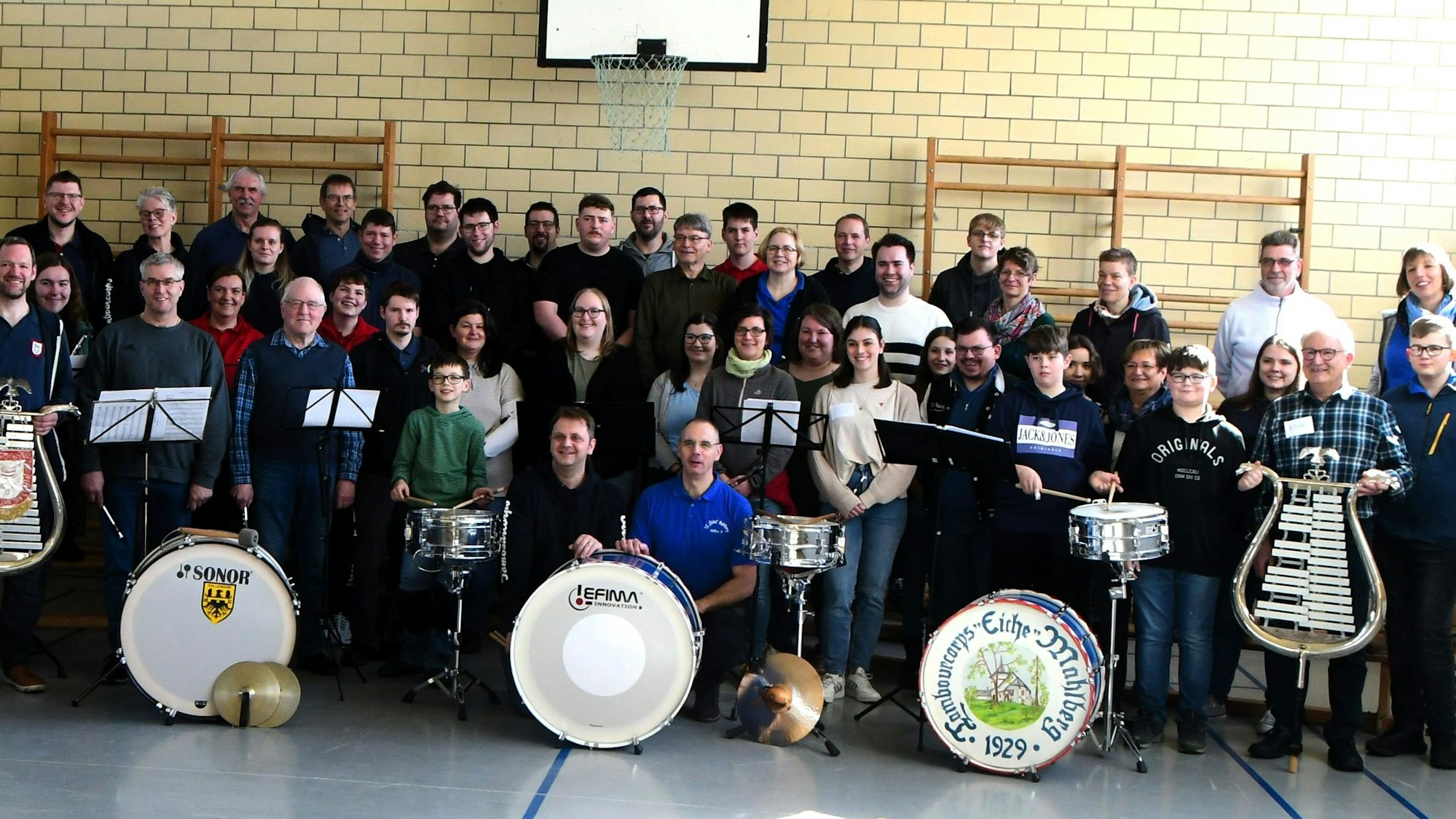 Zahlreiche Musiker stehen in einer Turnhalle. Einige halten Lyras, vor anderen stehen Pauken.