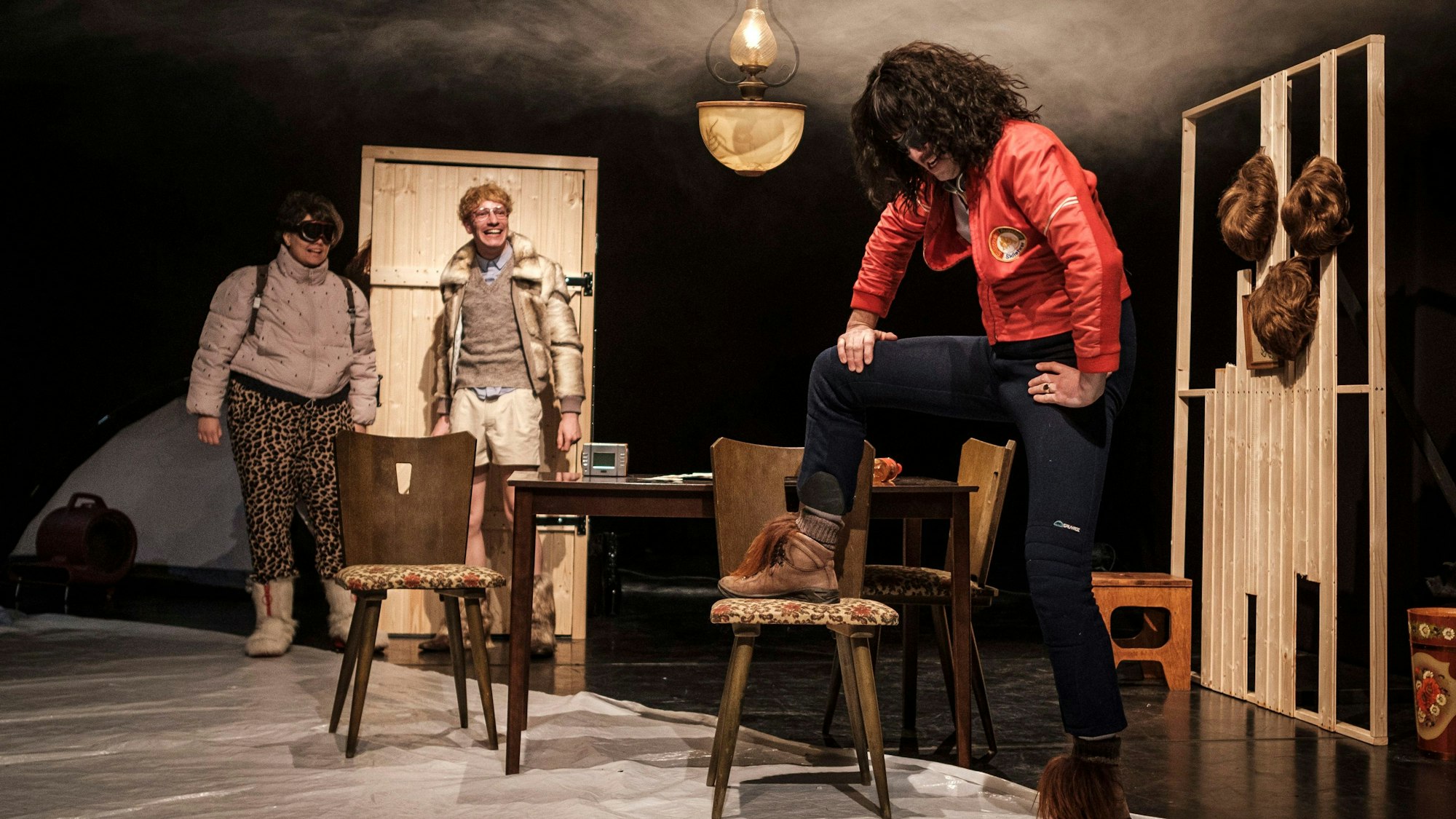 Im Vordergrund ist ein Schauspieler mit langen Haaren und einer roten Lederjacke, der energisch einen Fuß auf einem Stuhl abstützt, als würde er einen Berg besteigen. Die Bühne ist wie eine Almhütte eingerichtet. Im Hintergrund zwei weitere Bergsteiger in Winterkleidung.
