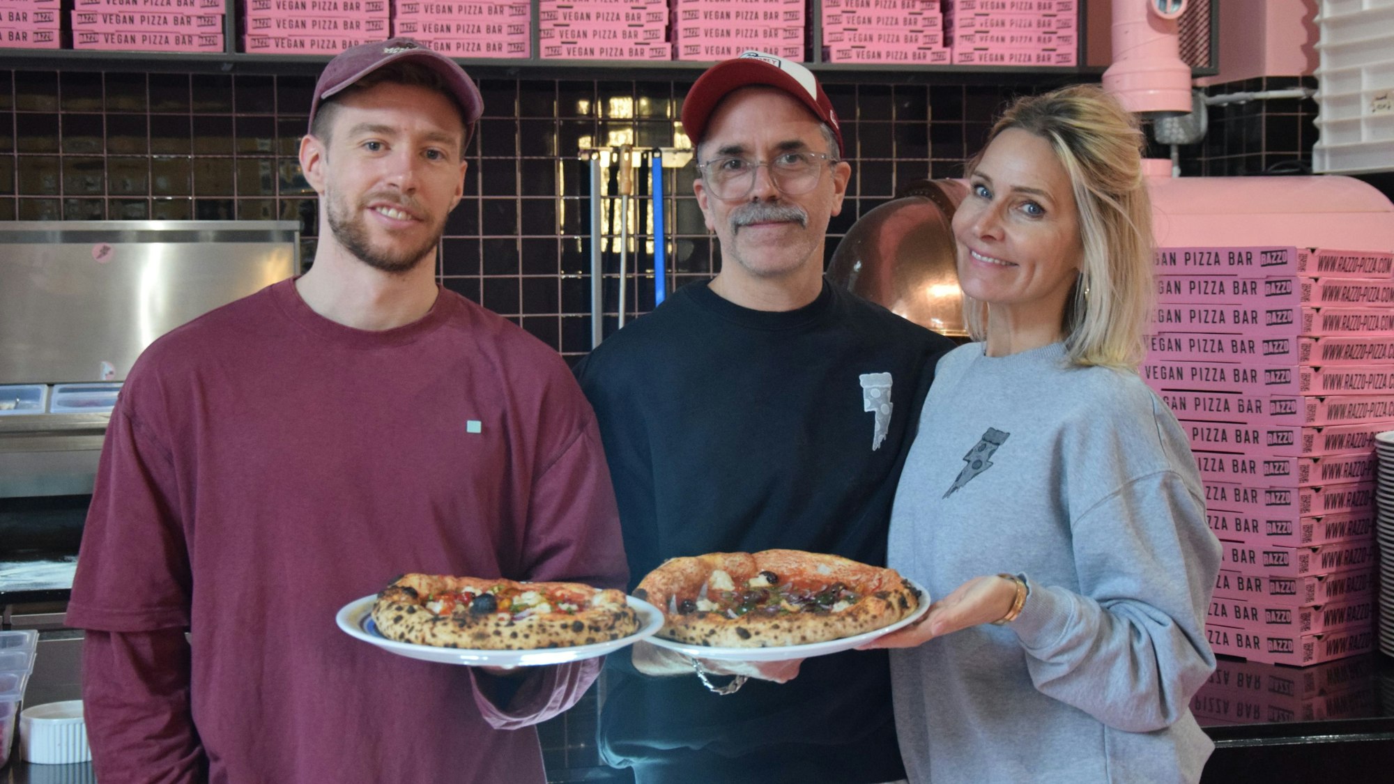 Zu sehen sind Mitchell Weiser, Ingo Noack und Verena Malta, die zwei Pizzen in die Kamera halten.