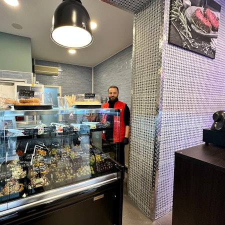 Ein Mann mit Bart und roter Jacke steht hinter einer Theke, in der Oliven und Frischkäse-Sorten kühl gehalten werden.