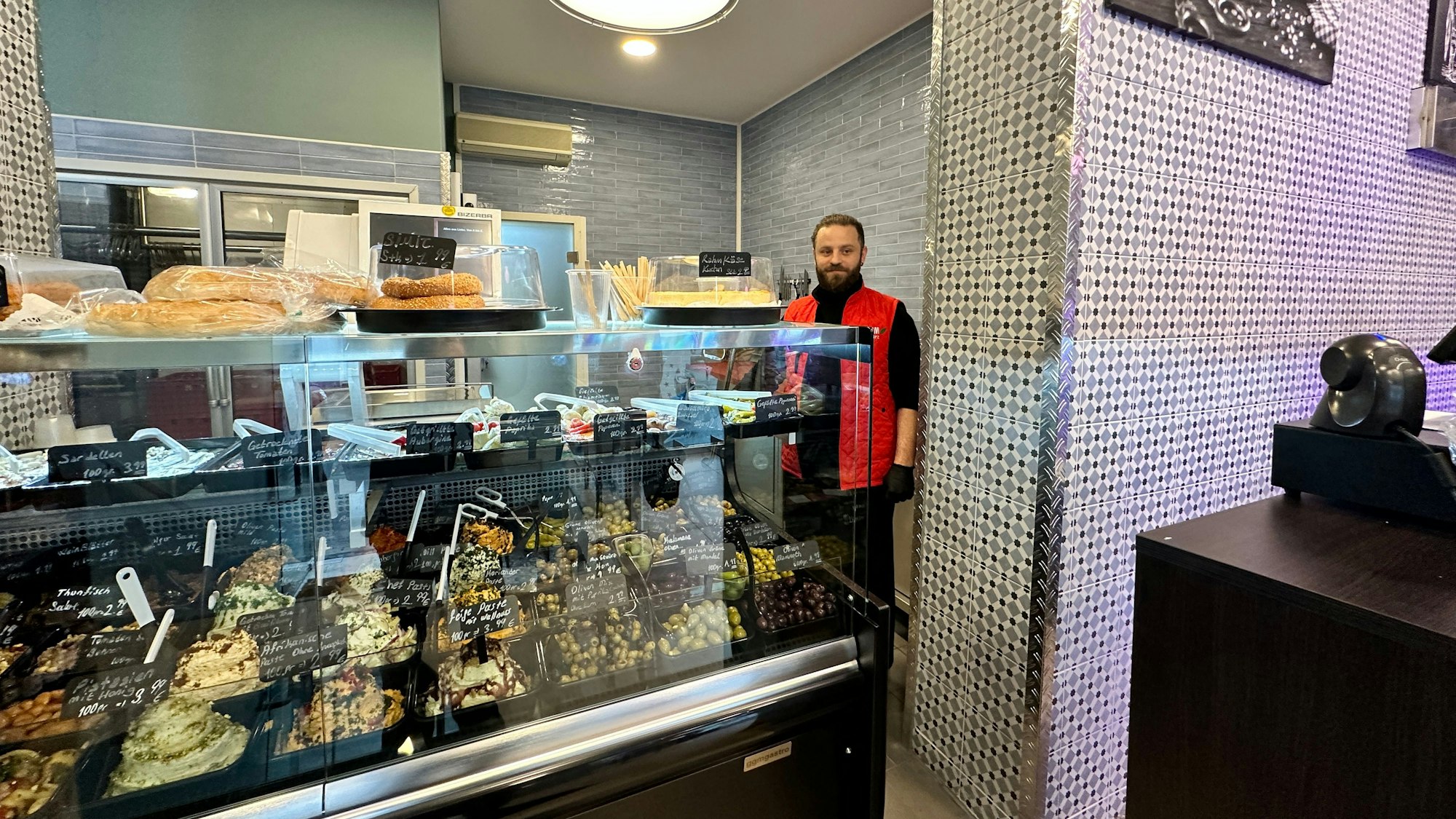 Ein Mann mit Bart und roter Jacke steht hinter einer Theke, in der Oliven und Frischkäse-Sorten kühl gehalten werden.