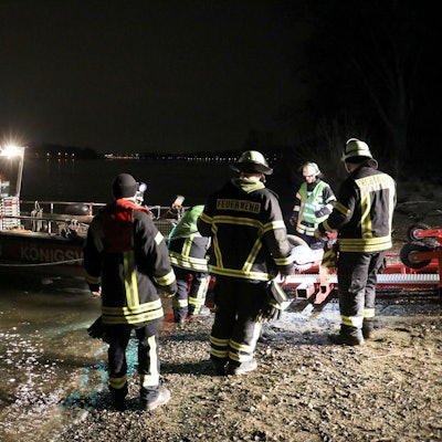 Feuerwehrleute stehen um ein Rettungsboot herum, das am Rheinufer halb auf dem Sand liegt.