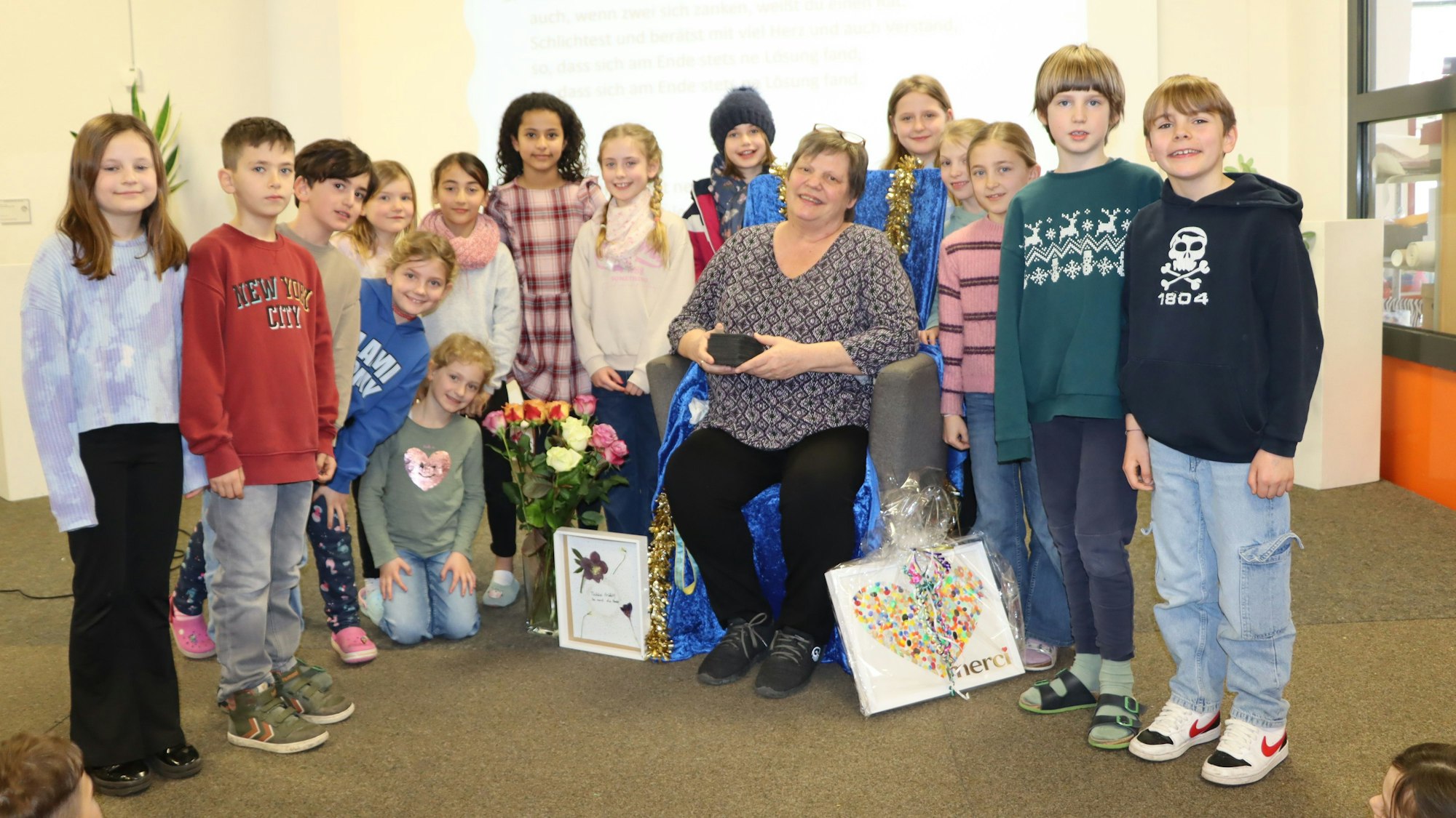 Eine Frau sitzt auf einem Sessel inmitten von Kindern mit Blumen und Präsenten.