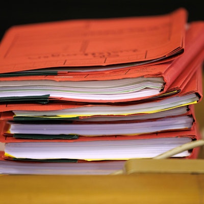 Mehrere rote Prozessakten liegen in einem Gerichtssaal zu Beginn eines Prozesses auf einem Tisch.