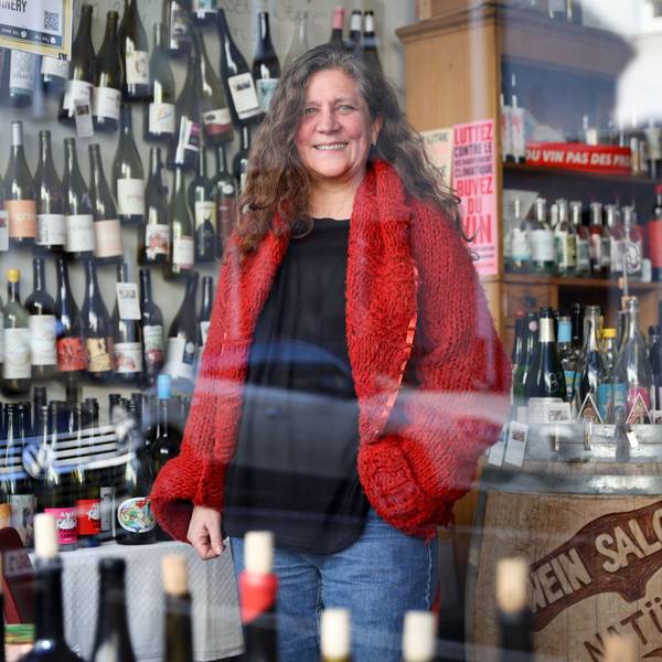 Eine Frau in roter Strickjacke und Jeans steht in einem Weinladen. Hinter ihr und auf einem großen Weinfass stehen und hängen zahlreiche Flaschen.