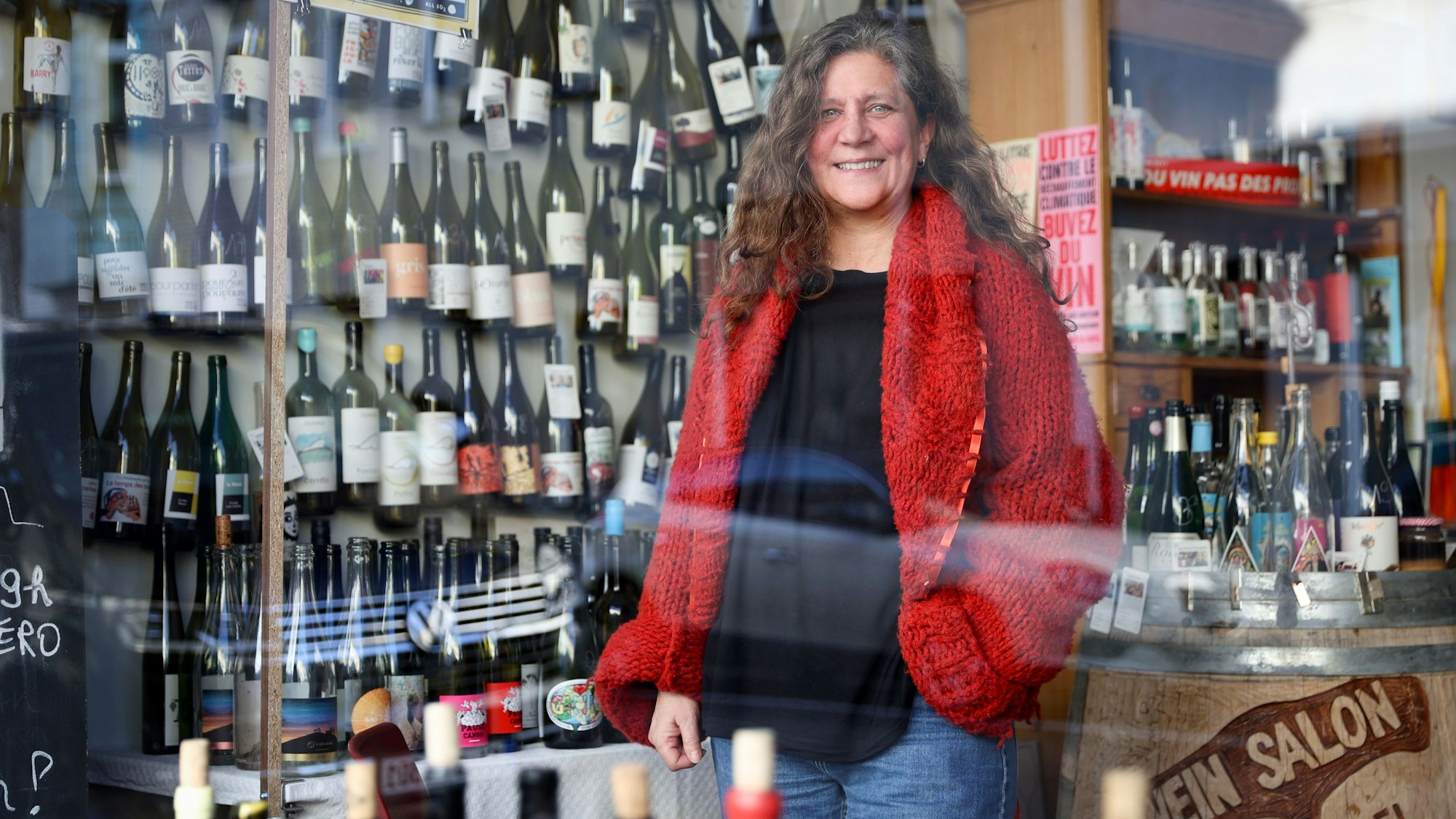 Eine Frau in roter Strickjacke und Jeans steht in einem Weinladen. Hinter ihr und auf einem großen Weinfass stehen und hängen zahlreiche Flaschen.