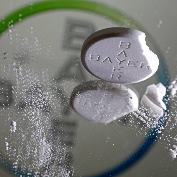 Eine Aspirin Tablette, an der eine Ecke abgebrochen ist, steht vor einem Bayer Logo auf einem Spiegel.