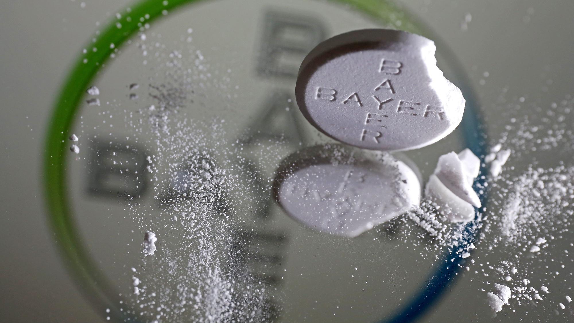 Eine Aspirin Tablette, an der eine Ecke abgebrochen ist, steht vor einem Bayer Logo auf einem Spiegel.