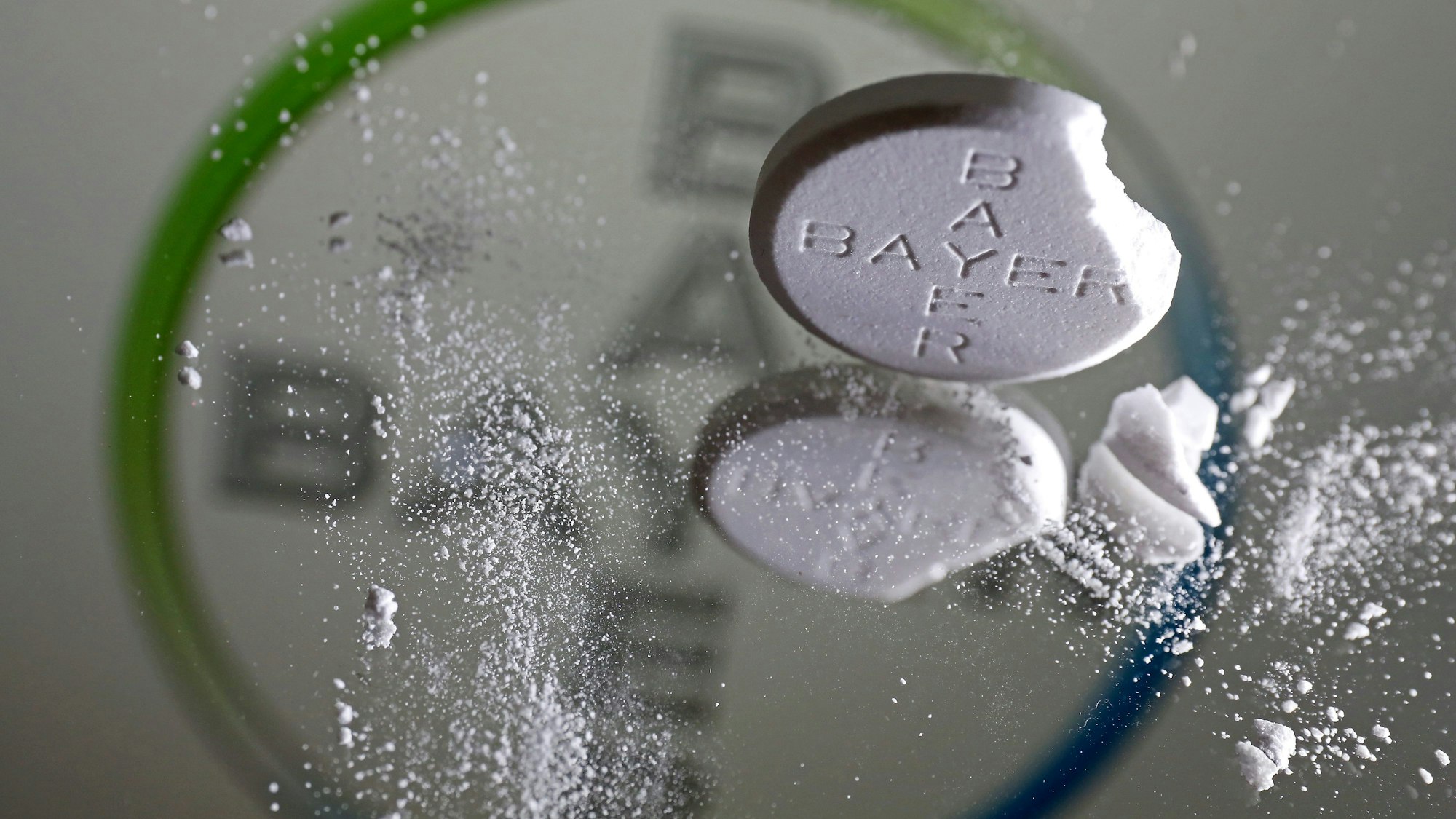 Eine Aspirin Tablette, an der eine Ecke abgebrochen ist, steht vor einem Bayer Logo auf einem Spiegel.