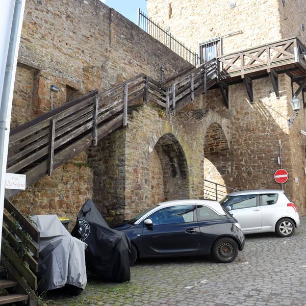 Das Bild zeigt die Holztreppe zum Wehrgang am Heisterbacher Tor aus Sicht der Werkbrücke. Vor der Treppe stehen Autos und Motorräder.