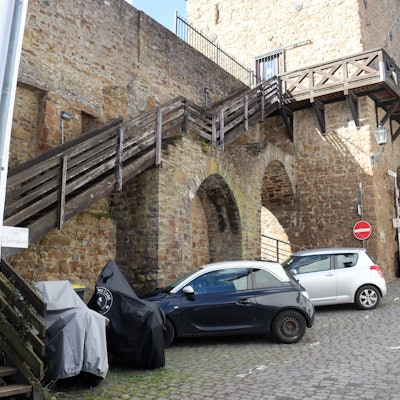 Das Bild zeigt die Holztreppe zum Wehrgang am Heisterbacher Tor aus Sicht der Werkbrücke. Vor der Treppe stehen Autos und Motorräder.