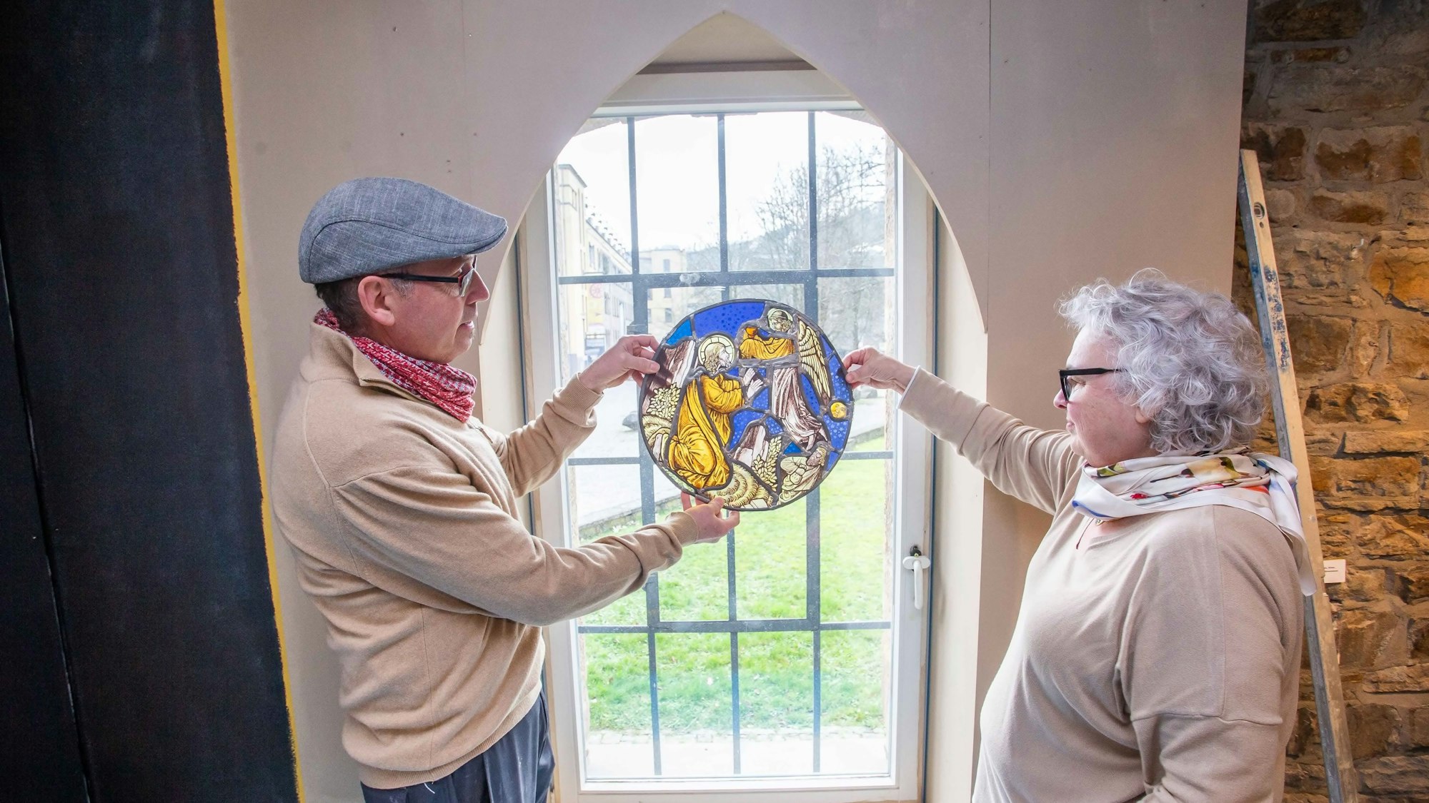 Ein Mann und eine Frau halten ein Glasmosaik in Händen, das Mosaik wird vom Tageslicht erhellt, das durch ein Fenster fällt.