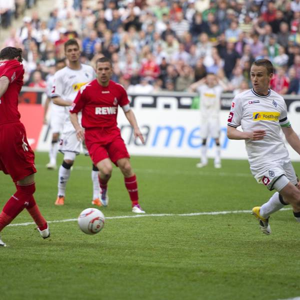 Der in rot gekleidete Stürmer des 1. FC Köln lässt zwei Gegenspieler aussteigen und erzielt ein Tor. Einer seiner Mitspieler schaut zu.