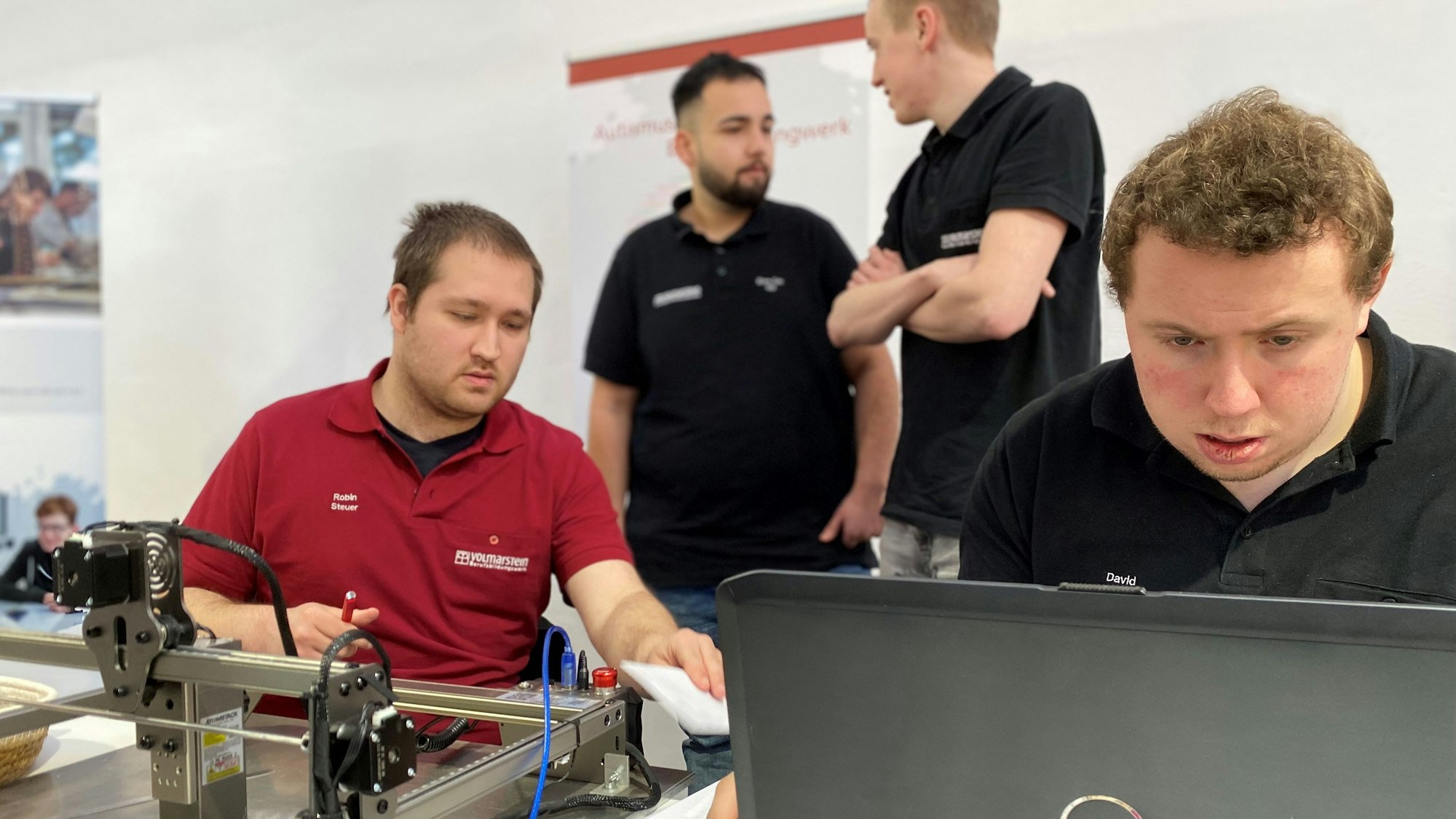 Ein junger Mann sitzt an einem Laptop, ein anderer an einer Lasermaschine. Im Hintergrund sind zwei weitere junge Männer zu erkennen.