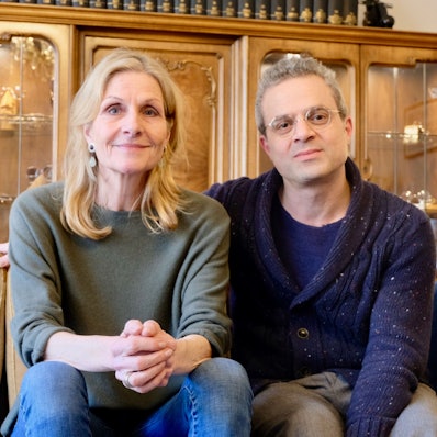 Eine Frau mit blondem Haar und ein Mann mit grauem Haar und Brille sitzen auf einem Sofa vor einer altmodischen Schrankwand.