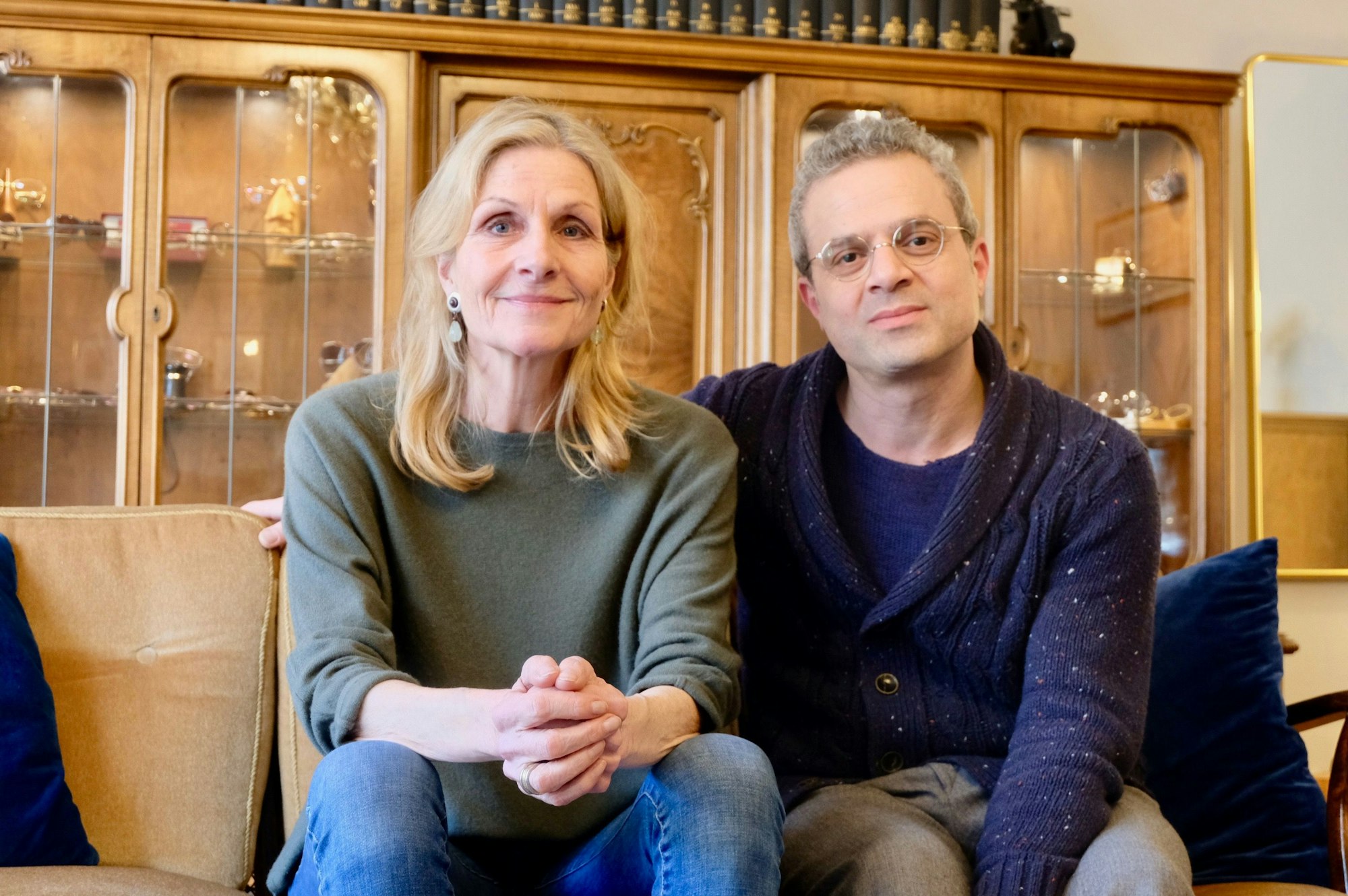 Eine Frau mit blondem Haar und ein Mann mit grauem Haar und Brille sitzen auf einem Sofa vor einer altmodischen Schrankwand.