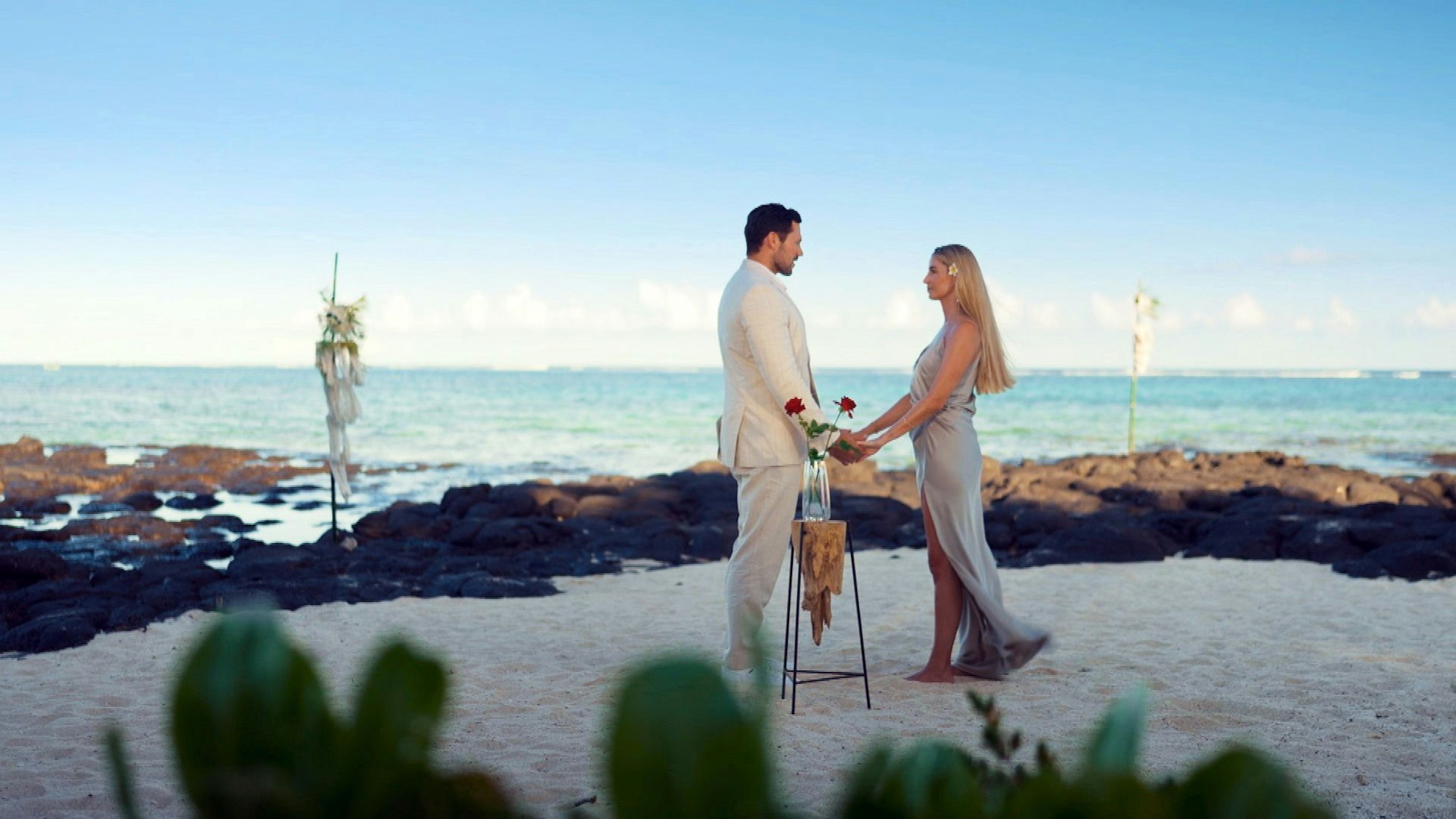 Larissa und Sebastian stehen gemeinsam an einem Strand und halten Händchen.