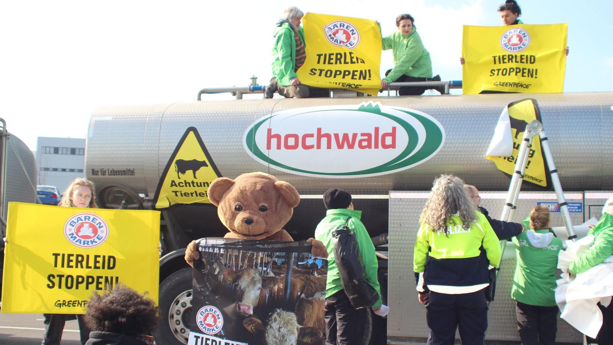 Mehrere Demonstranten stehen vor und sitzen auf einem Lkw, auf dem das „Hochwald“-Logo angebracht ist. Sie halten Plakate mit der Aufschrift „Tierleid stoppen“ hoch, eine Demonstrantin trägt ein Bärenkostüm, wie es aus der früheren Werbung von „Bärenmarke“ bekannt ist.