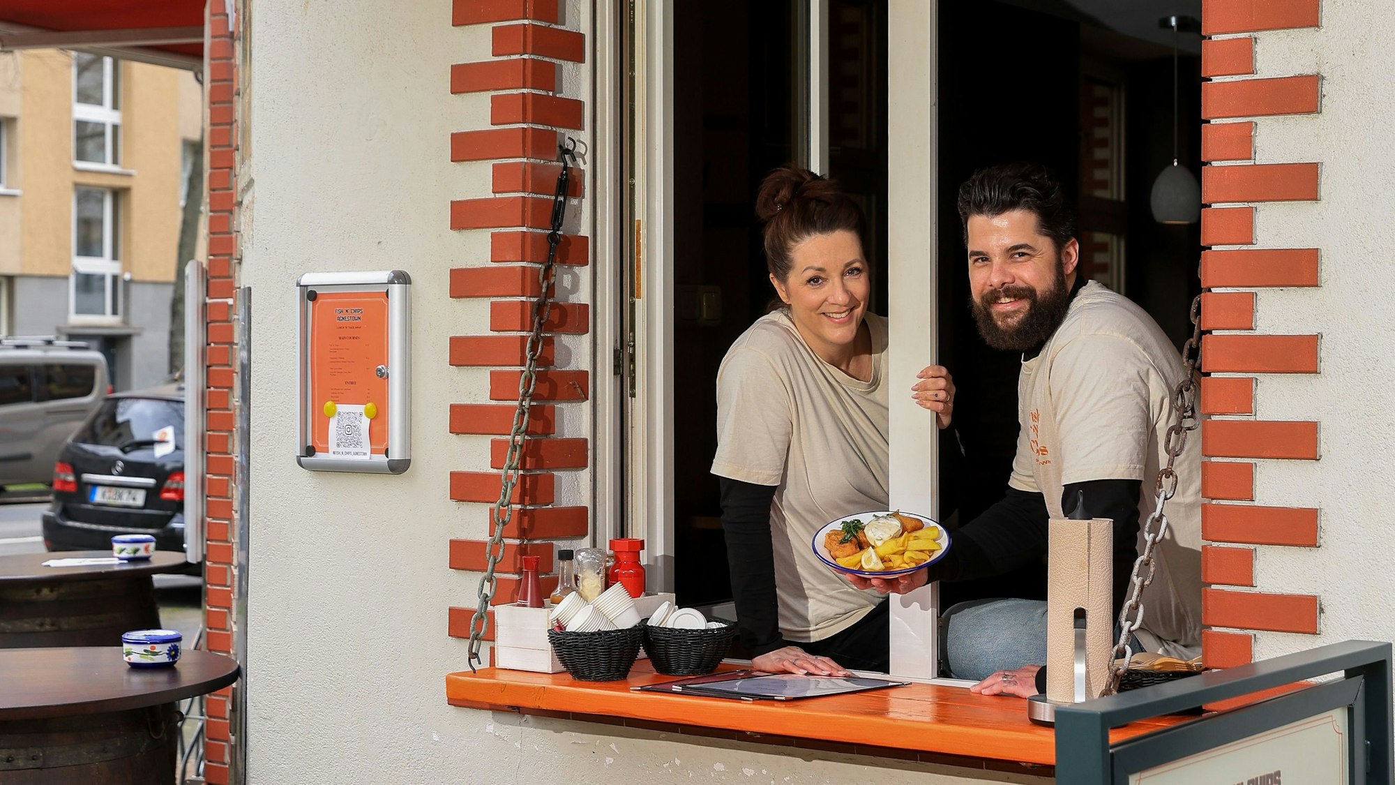 Eine Frau und ein Mann sitzen in einem Verkaufsfenster, der Mann hält einen Teller mit Fish and Chips in der Hand.