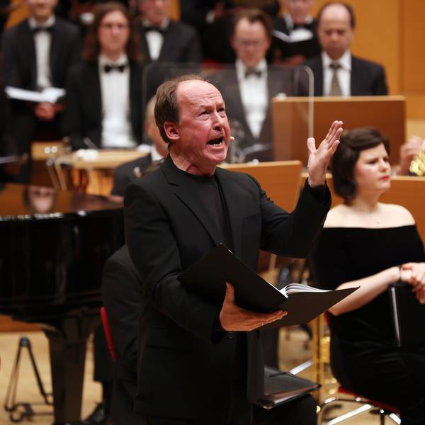 Ulrich Noethen hält eine offene Mappe in der rechten und gestikuliert mit der linken Hand. Er trägt einen schwarzen Anzug. Im Hintergrund das Orchester.