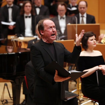 Ulrich Noethen hält eine offene Mappe in der rechten und gestikuliert mit der linken Hand. Er trägt einen schwarzen Anzug. Im Hintergrund das Orchester.