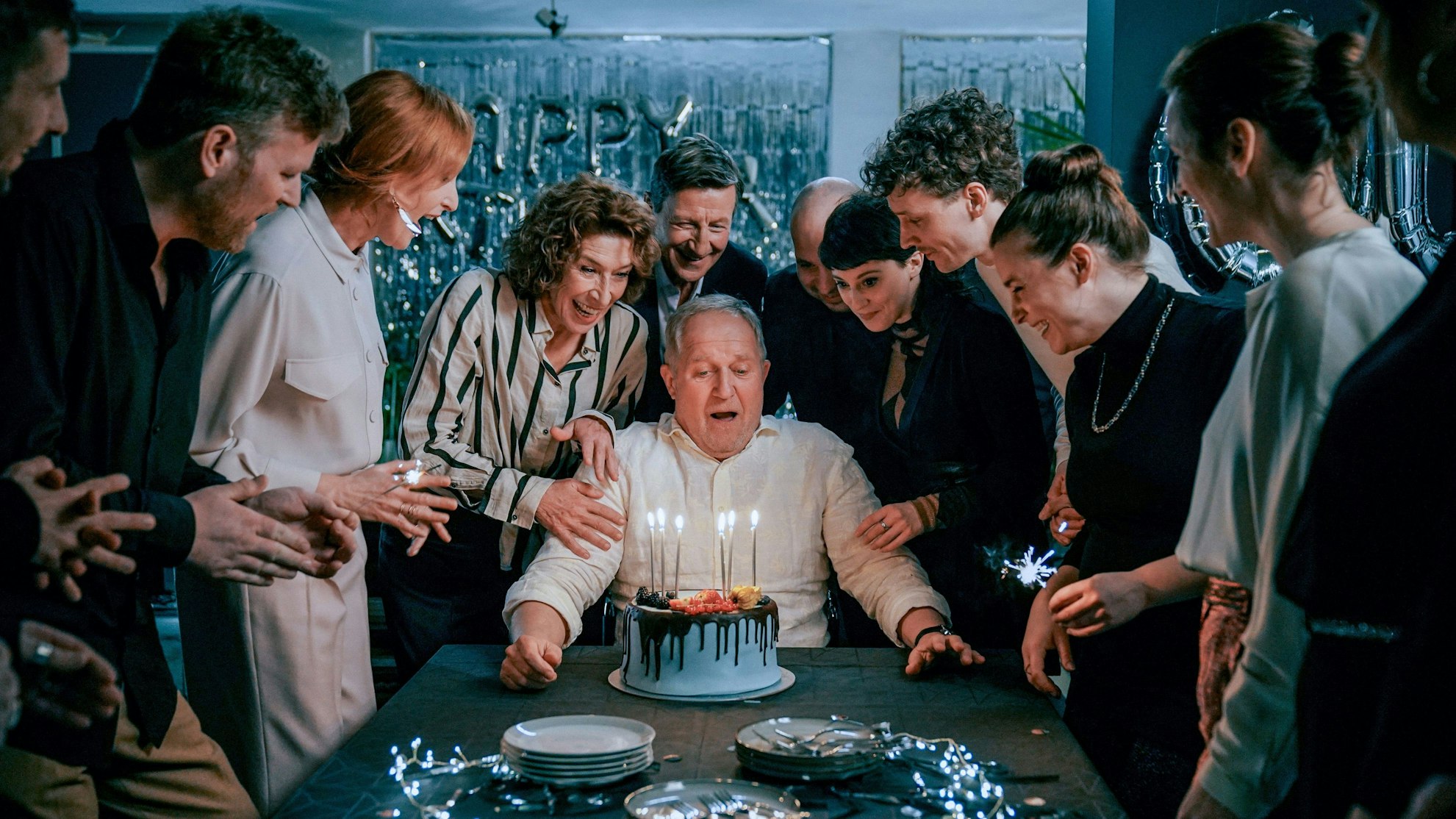 Moritz Eisner sitzt am Tisch vor einer Torte mit Kerzen. Um ihn herum steht die feiernde Gesellschaft, darunter auch seine Partnerin Bibi Fellner.