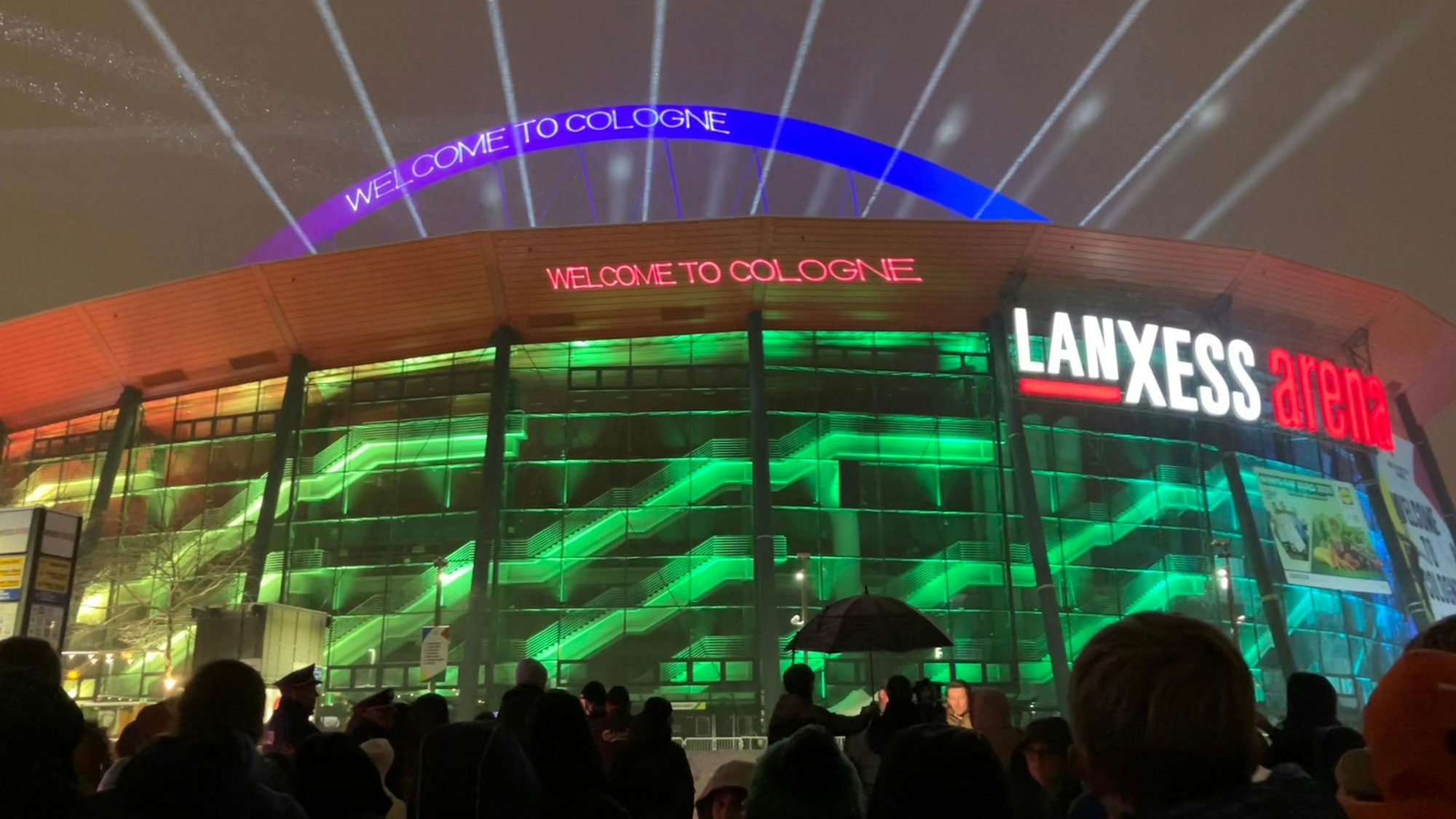 Menschen stehen vor der beleuchteten Lanxess-Arena, die den Schriftzug „Welcome to Cologne“ trägt.